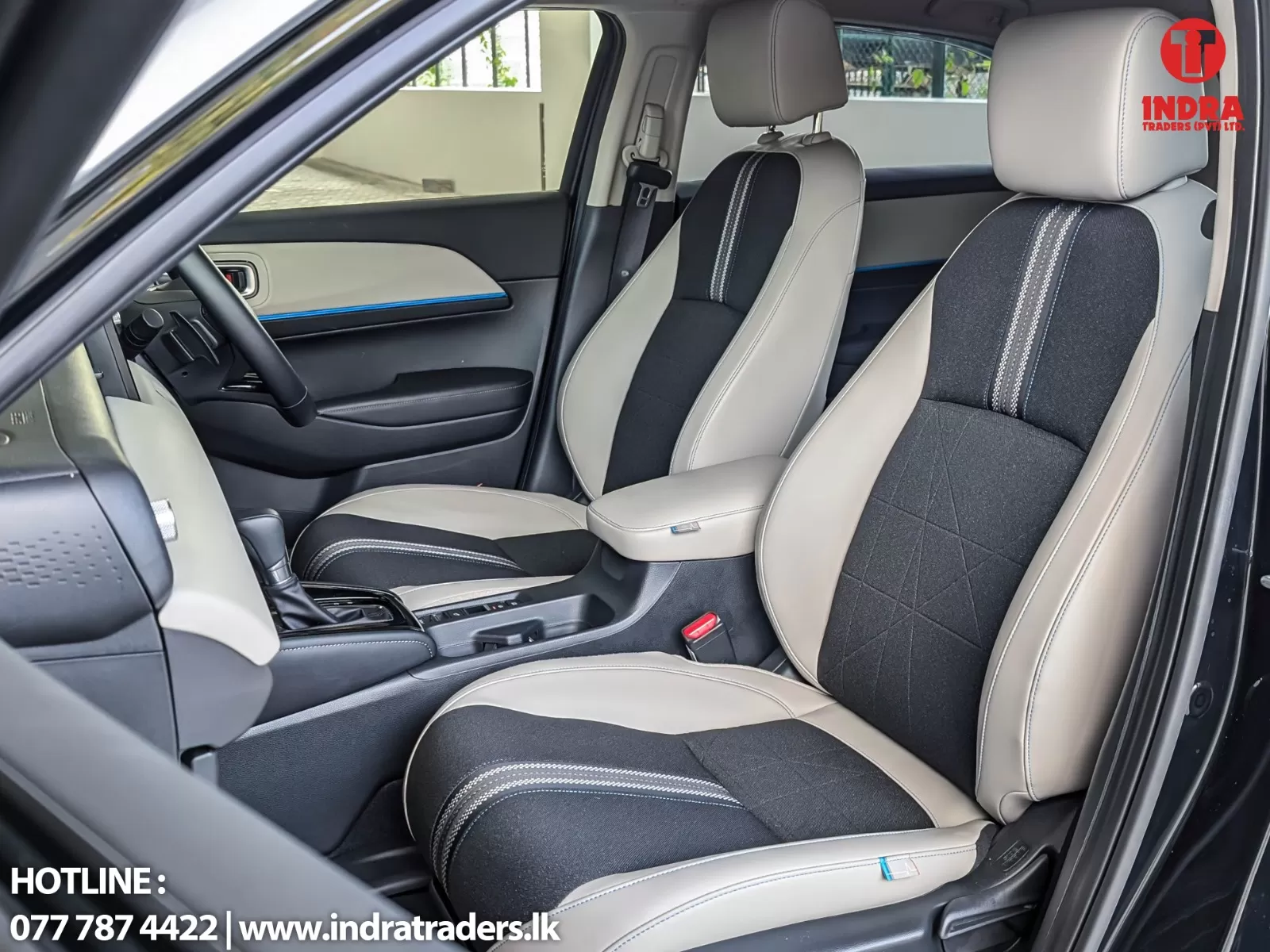 Front interior of a car with beige and black two-tone seats, center console, and a visible Indra Traders logo in the top-right corner.