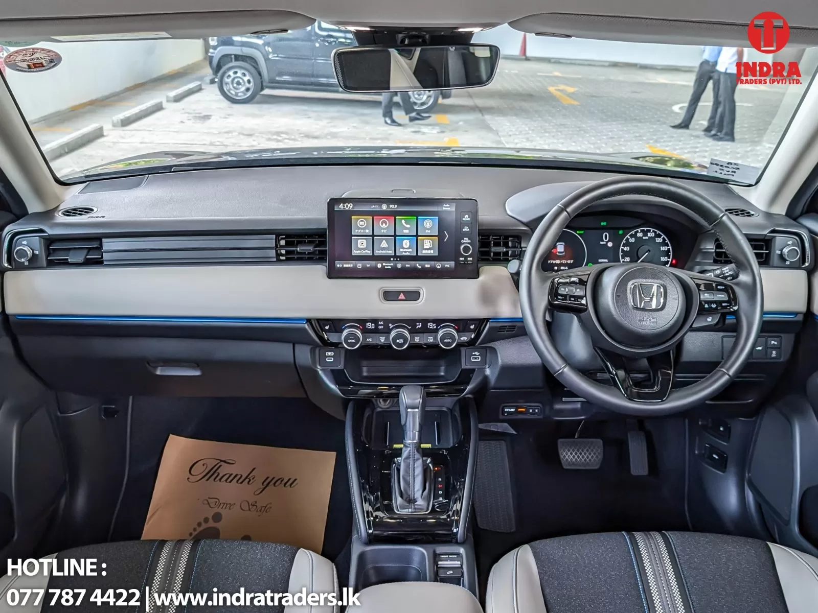 Inside view of a Honda car cockpit: steering wheel, digital instrument cluster, center touchscreen, climate controls, gear shifter, and a 'Thank you' mat on the floor.