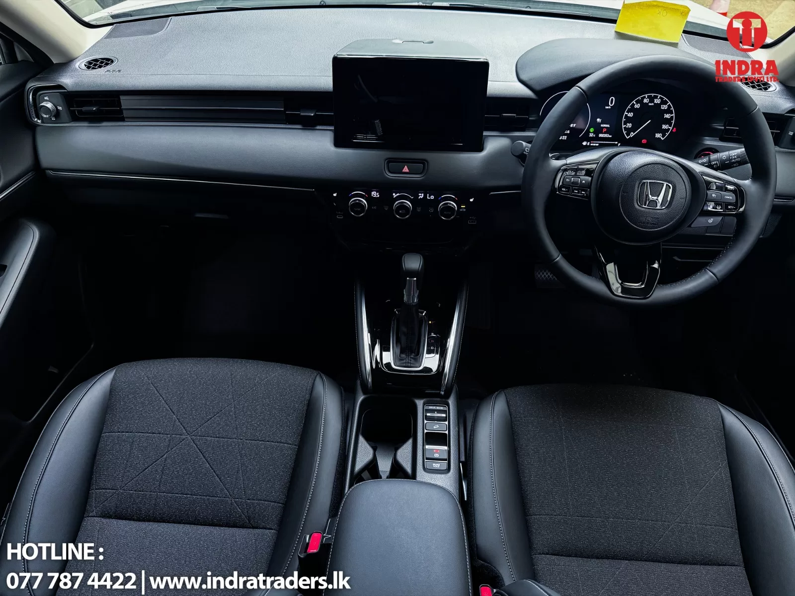 Interior of a Honda car showing the dashboard, large central touchscreen, climate control knobs, steering wheel with controls, and gear shifter between two front seats; Indra Traders logo in the top-right and hotline text in the bottom-left.