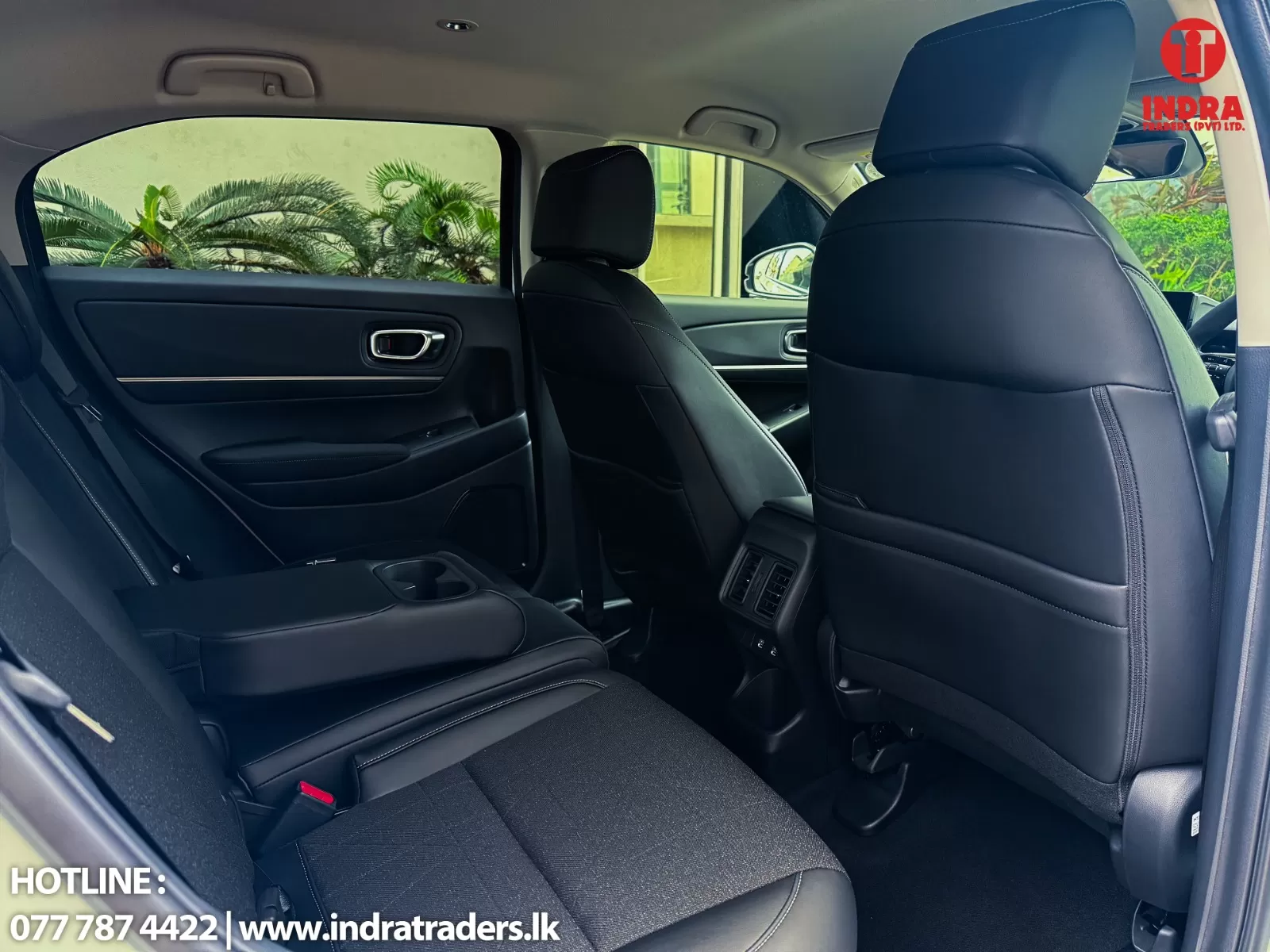 Backseat of a black leather SUV with two rows of seats; greenery visible through the side window; Indra Traders logo in the top-right and hotline text at the bottom left.