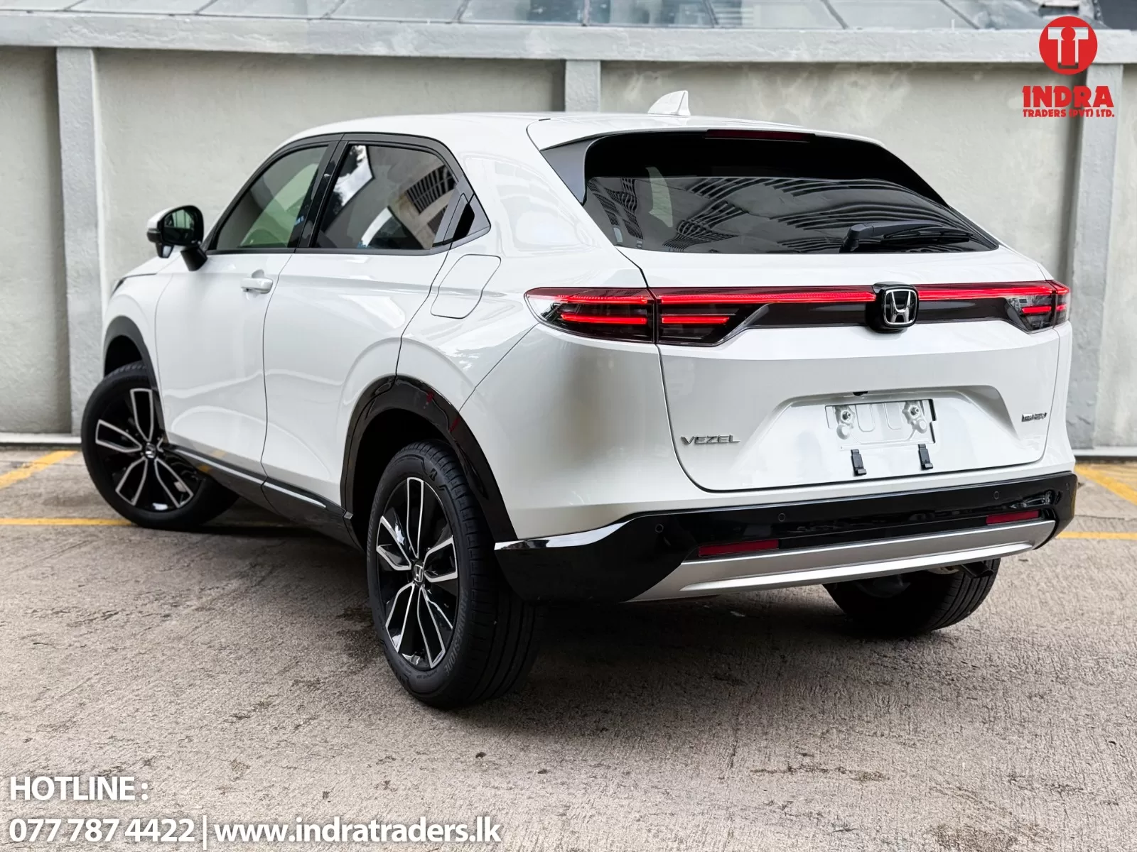 Rear three-quarter view of a white Honda Vezel SUV in a parking lot, showing red taillights and black rear spoiler with alloy wheels; dealership watermark top-right.