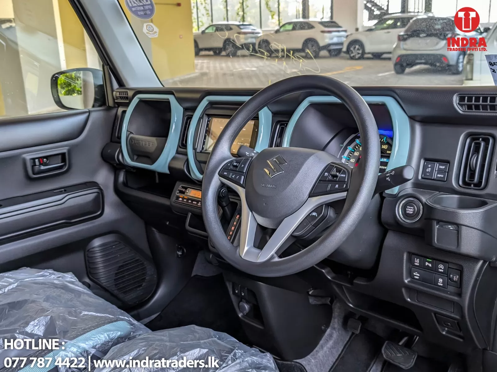 Interior of a car cabin showing the steering wheel, dashboard, and center touchscreen with turquoise accents; truck-like gear lever and control buttons visible; Indra Traders logo in top-right corner and contact info at bottom-left.
