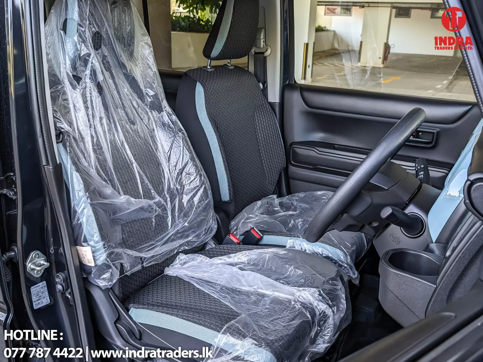 Front seat area of a car with plastic protective covers on the driver and passenger seats; steering wheel visible inside a showroom setting; Indra Traders logo in the top-right corner.