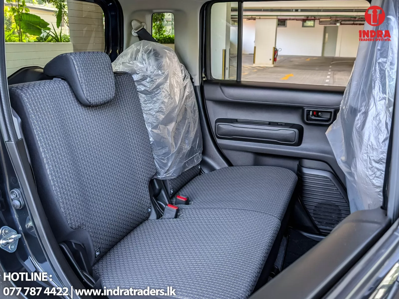Back seat of a car with gray patterned upholstery; the front seat is covered in plastic, view of a parking garage through the window, Indra Traders watermark in corner.