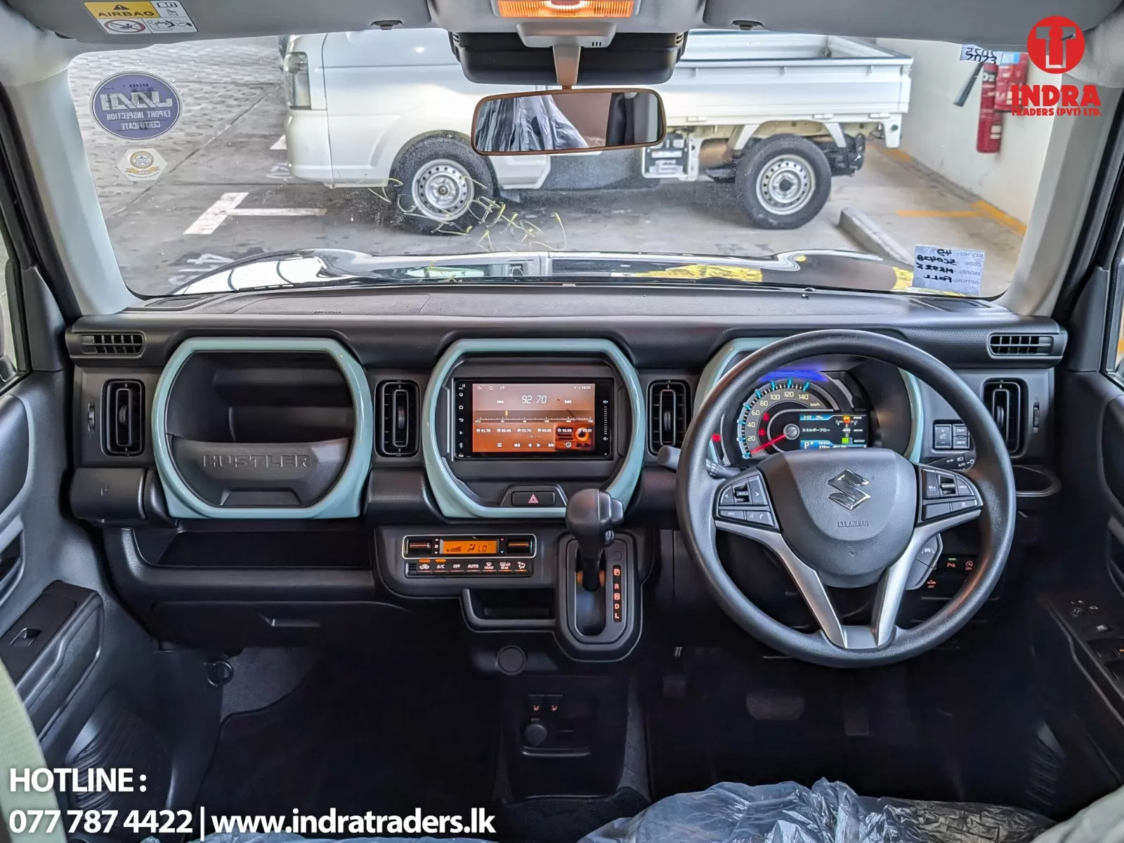 Interior view of a right-hand-drive SUV dashboard: steering wheel with Suzuki emblem, central touchscreen, climate controls, and gear selector, with a white pickup truck visible through the windshield outside.