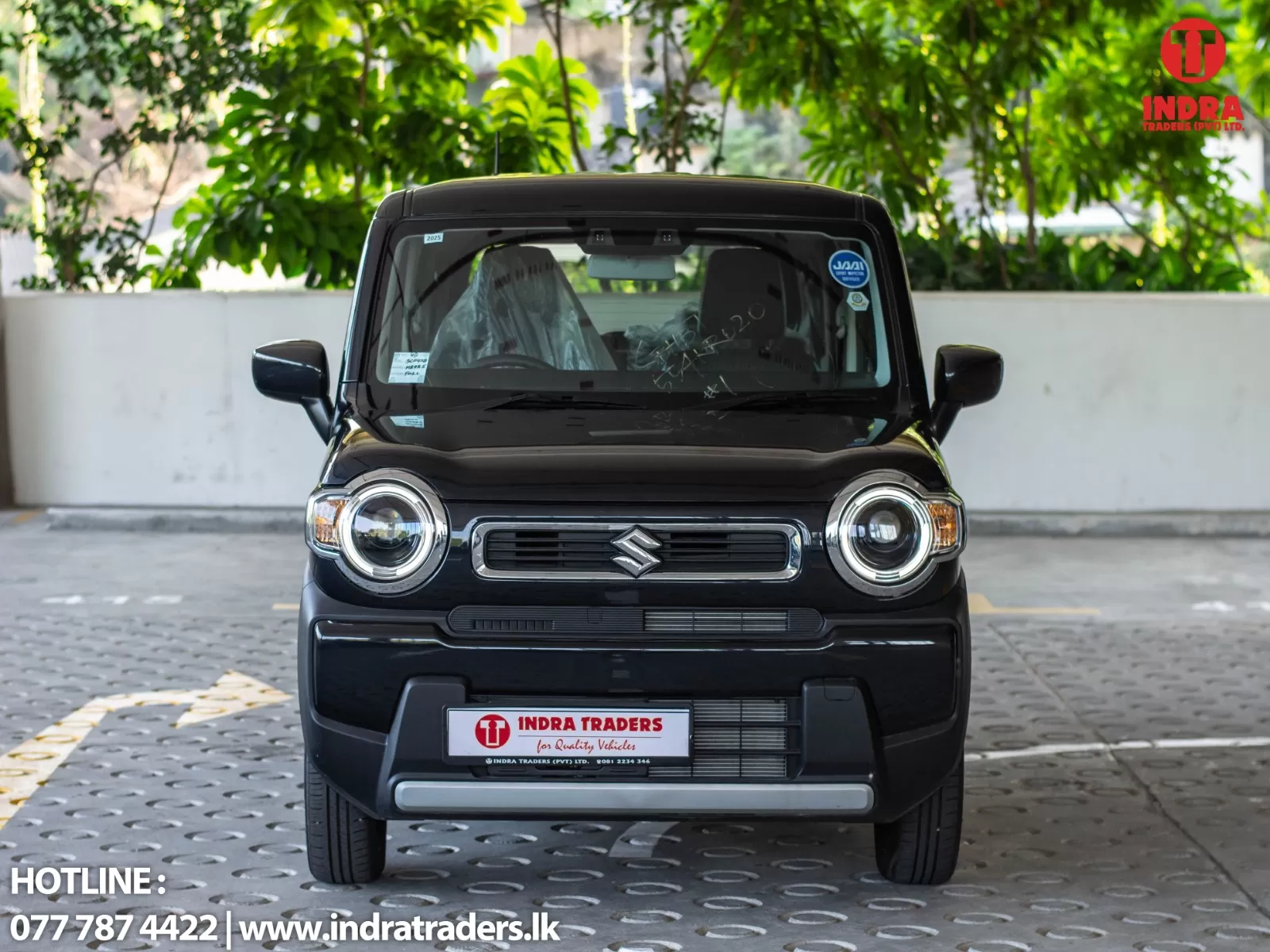 Front view of a black Suzuki SUV parked in a dealership lot, with the INDRA TRADERS logo in the top-right and contact text at the bottom left.