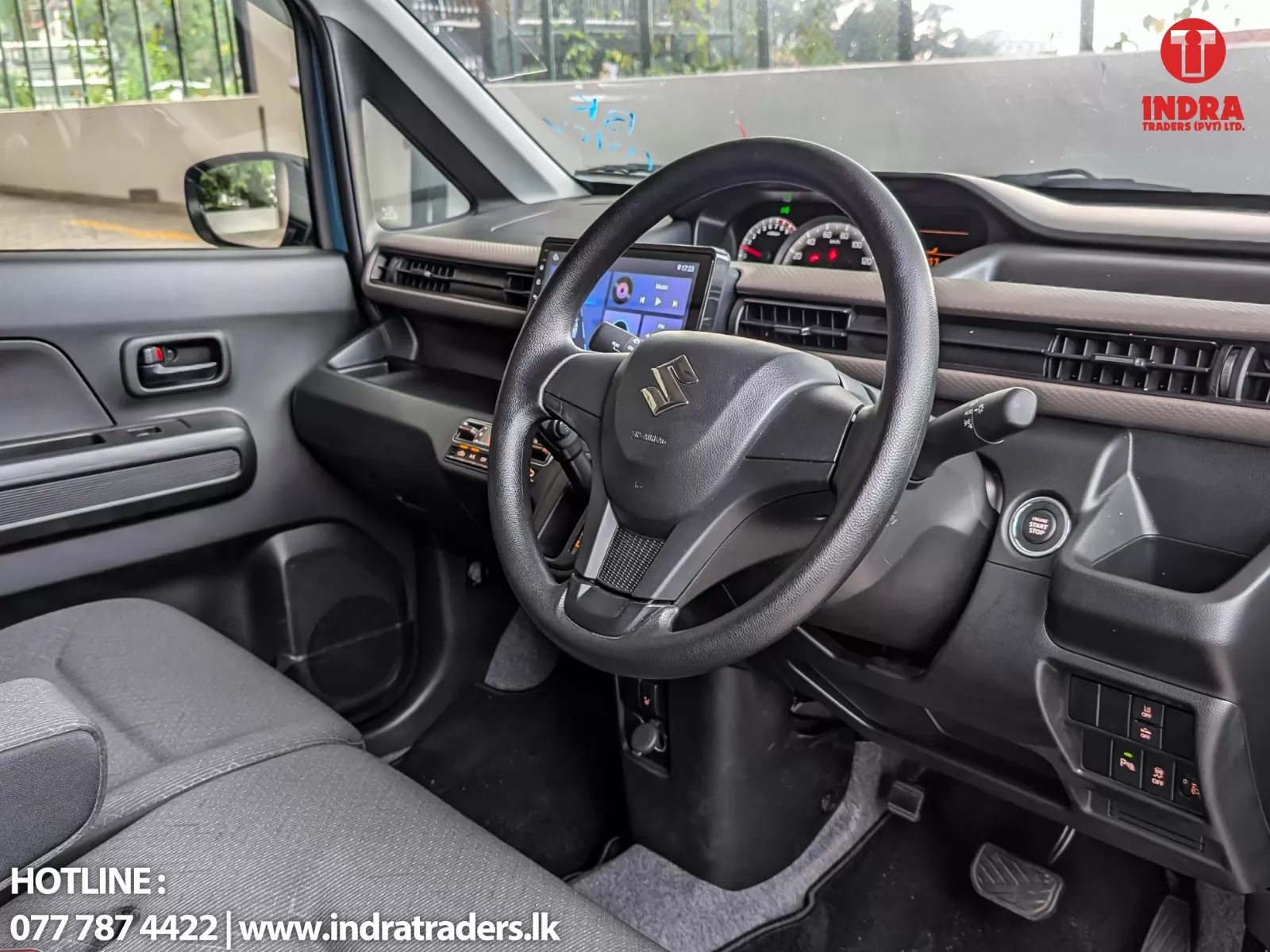 Interior view from the driver’s seat showing a steering wheel with the Suzuki emblem, touchscreen infotainment, dashboard gauges, push‑start button, and an Indra Traders logo on the windshield.