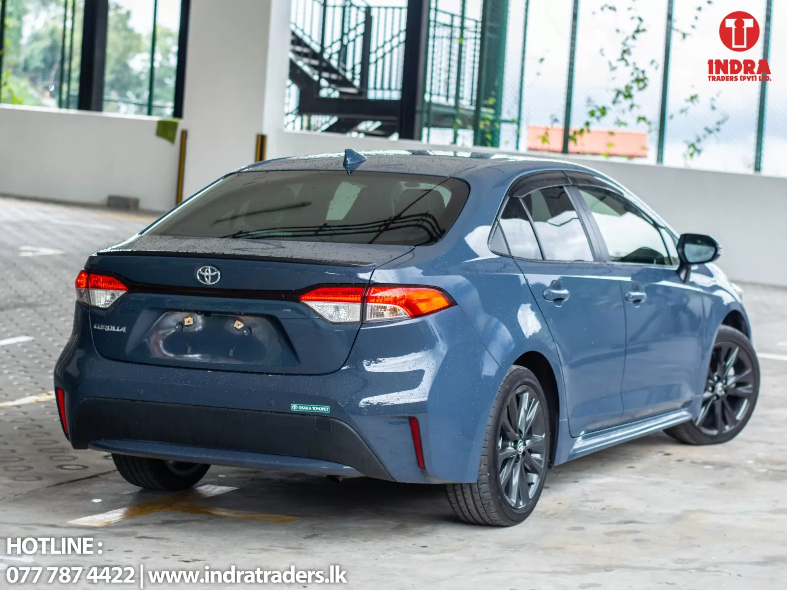 Blue Toyota sedan parked in a covered lot with a white wall and logo in the top-right corner (Indra Traders).