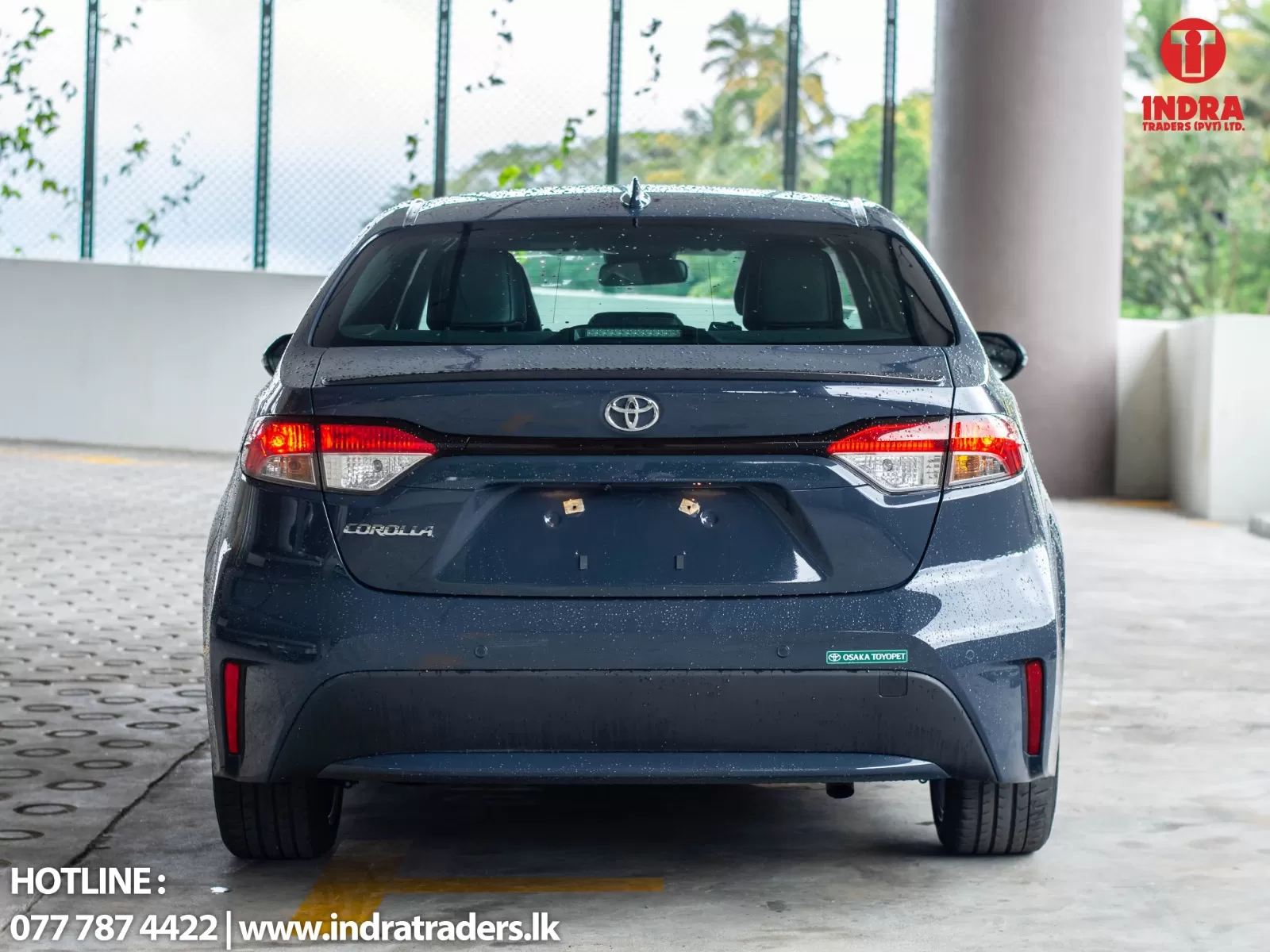 Rear view of a dark blue Toyota Corolla in a showroom/parking area, with Indra Traders logo in the top-right corner and contact info at the bottom-left.