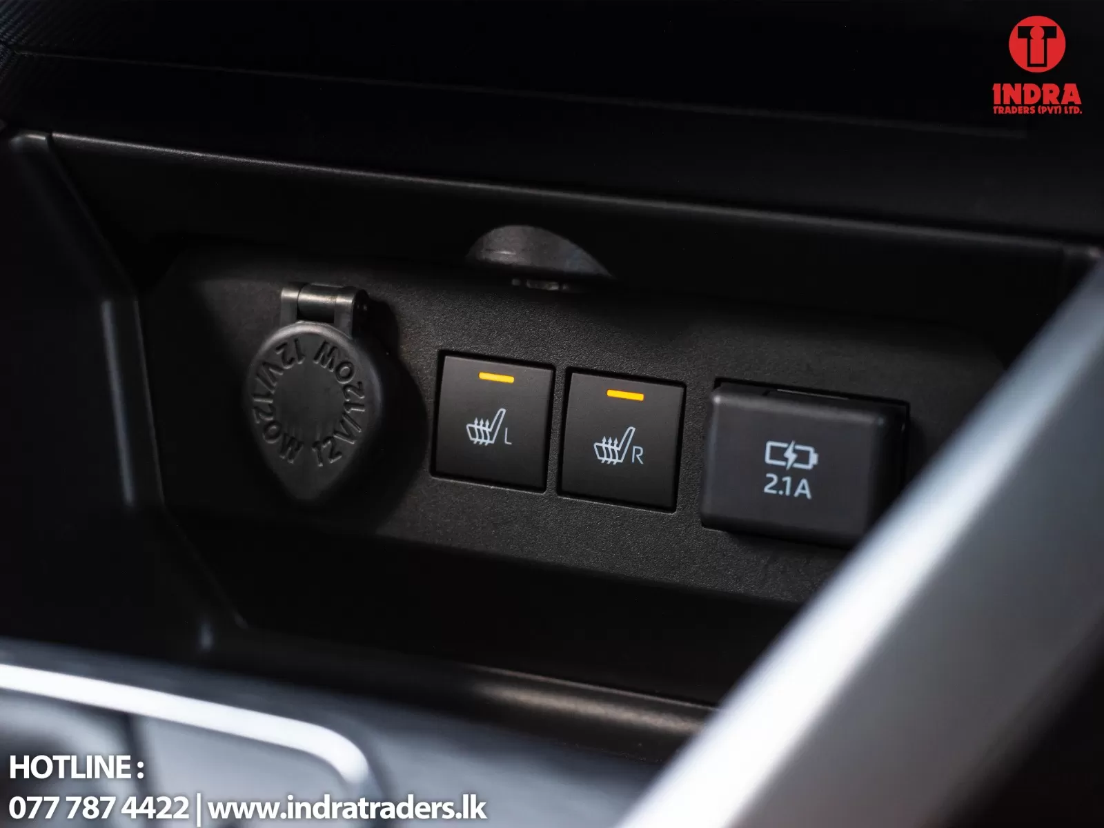 Close-up of car center console: left 12V power port cap, two heated-seat buttons (L and R) with orange indicators, and a 2.1A USB/C ports panel with logo in corner.