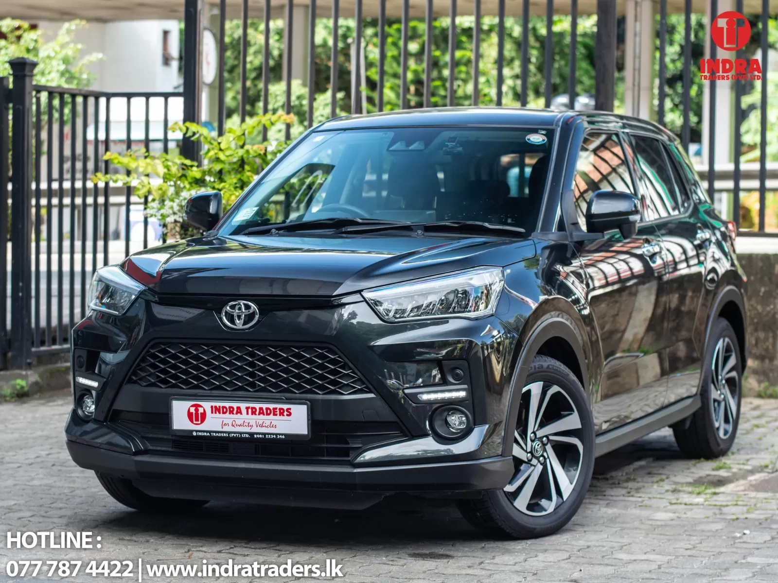 Black Toyota SUV parked outdoors near a fence, showing the Indra Traders logo on the top-right and the dealership hotline at the bottom.