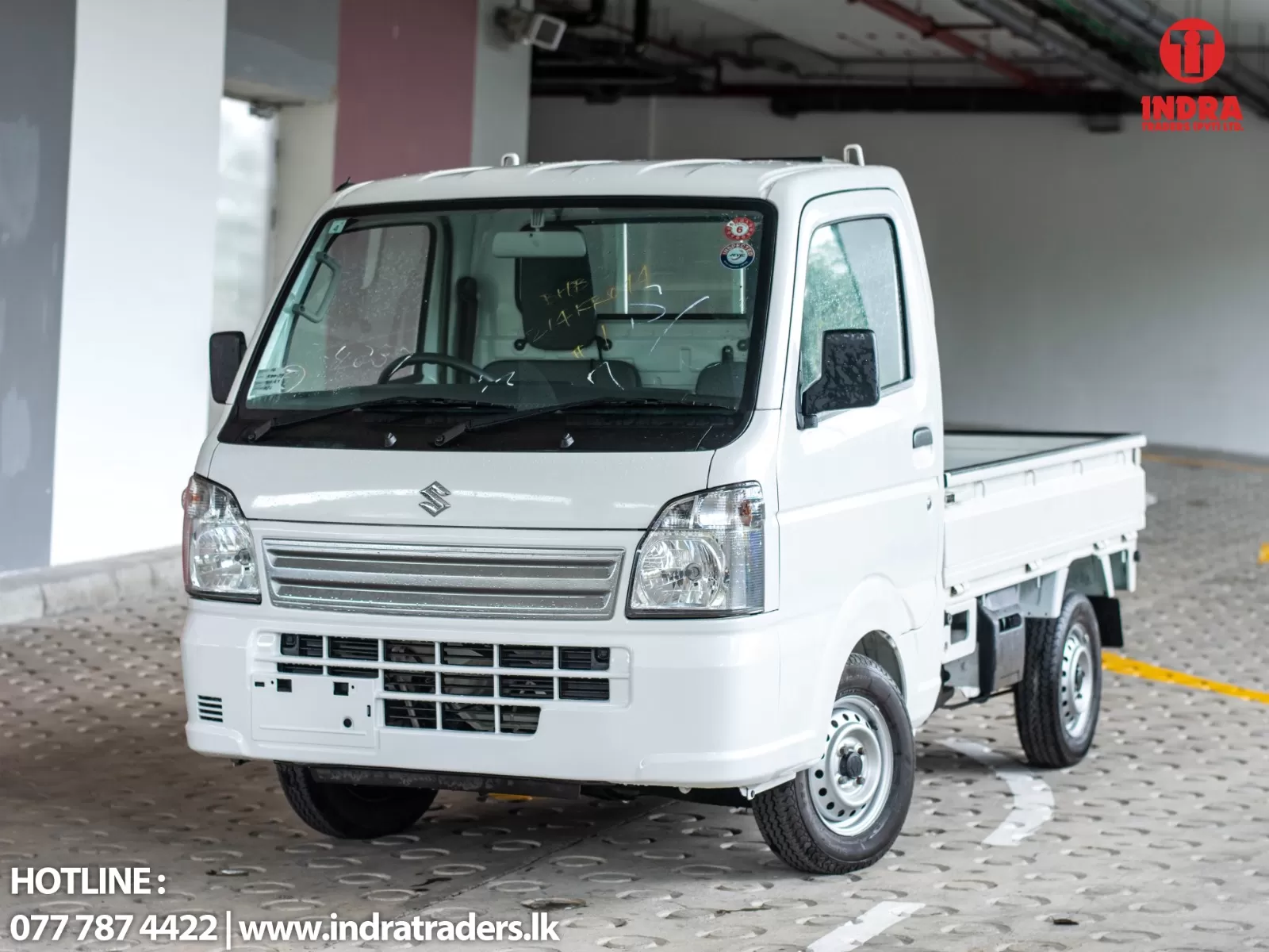 White Suzuki mini pickup in a showroom, front three-quarter view with Indra Traders logo in the top-right corner and contact text at the bottom.