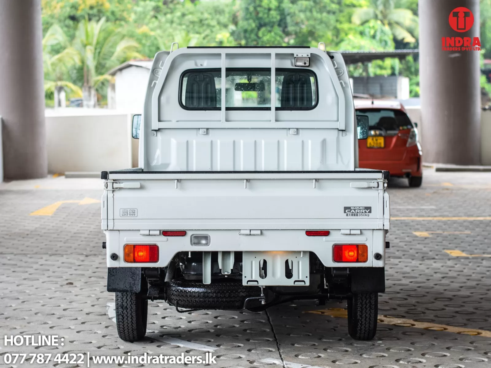 Rear view of a white Suzuki Carry pickup in a covered parking area, with the Indra Traders logo in the top-right corner and contact info at the bottom-left.