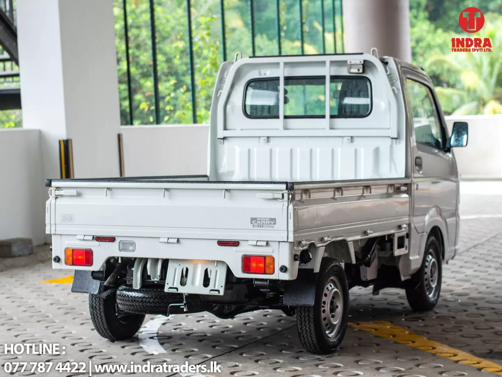 White Suzuki Carry pickup in a showroom, with INDRA Traders logo in the top-right and hotline text at bottom-left.
