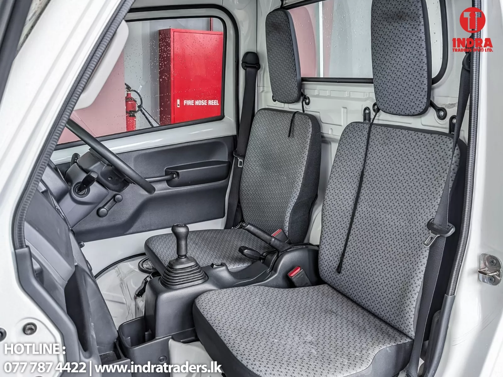 Interior of a light-gray truck cab with two front seats, a gear stick, and a steering wheel; a red fire hose reel is visible through the window outside.