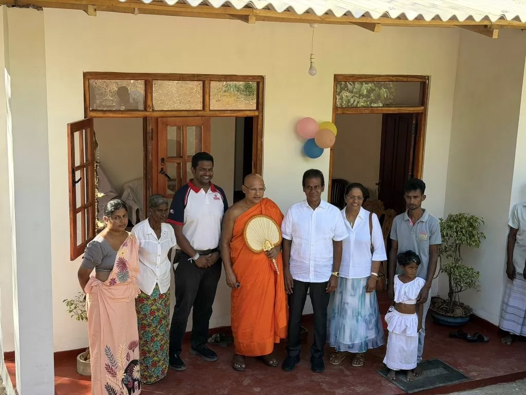 Group of people posing on a porch for a celebration; a Buddhist monk in orange robes holds a fan beside others.