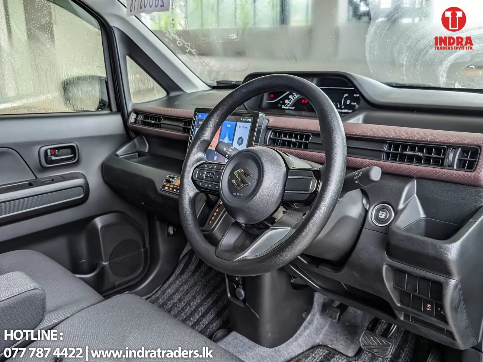 Car interior showing steering wheel with logo, central touchscreen, and dashboard on a rainy day; Indra Traders watermark in top-right corner.