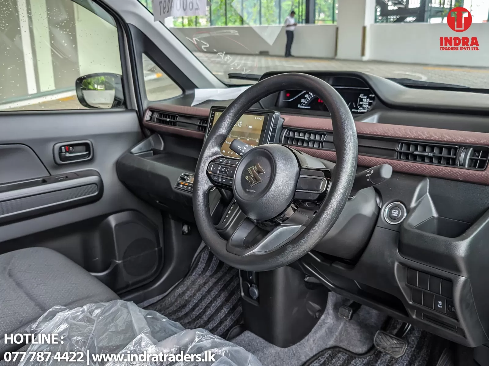 Car interior: steering wheel with Suzuki logo, touchscreen display, brown dashboard trim, push-start button and center console visible from the driver’s seat. Outside through the windshield, a person stands near a white wall.