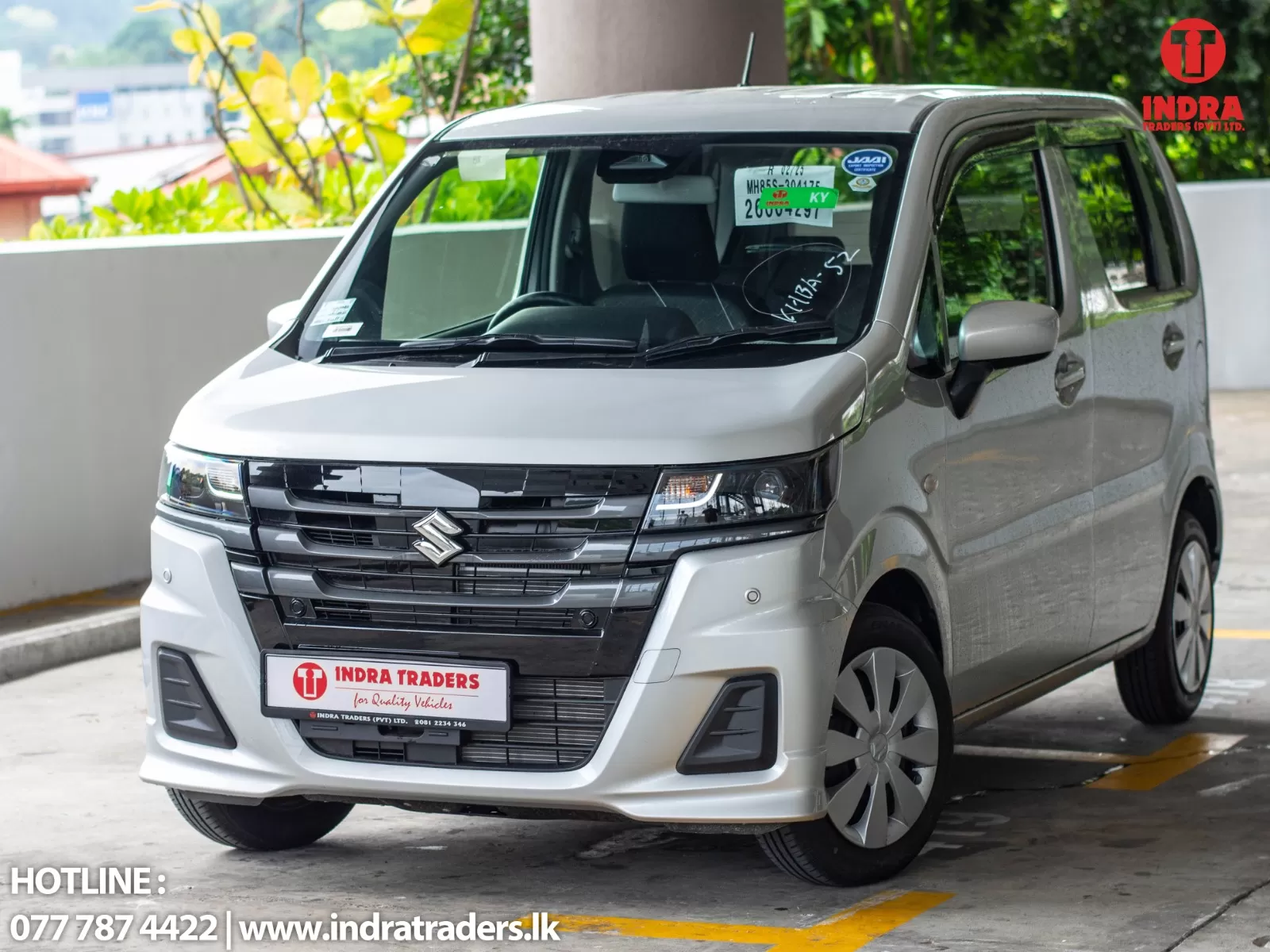 Front three-quarter view of a silver Suzuki van parked in a lot, with Indra Traders branding on the windshield and the hotline/contact text at the bottom left; logo visible in the top-right corner.