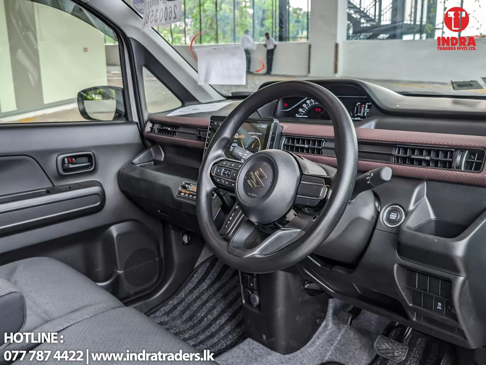 Front interior of a Suzuki car showing steering wheel, digital instrument cluster, center touchscreen, and push-button start.