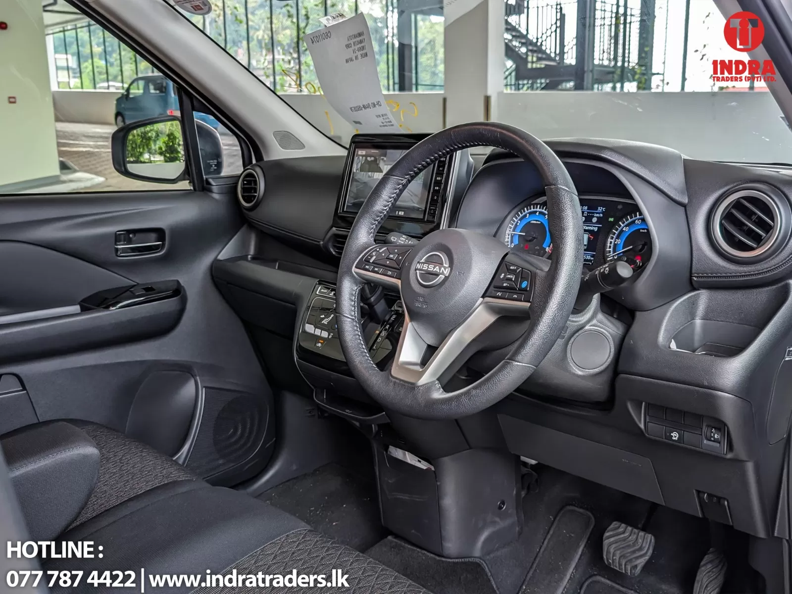 Interior of a Nissan vehicle showing the steering wheel, center console, and instrument cluster; contact text/logo in corner.