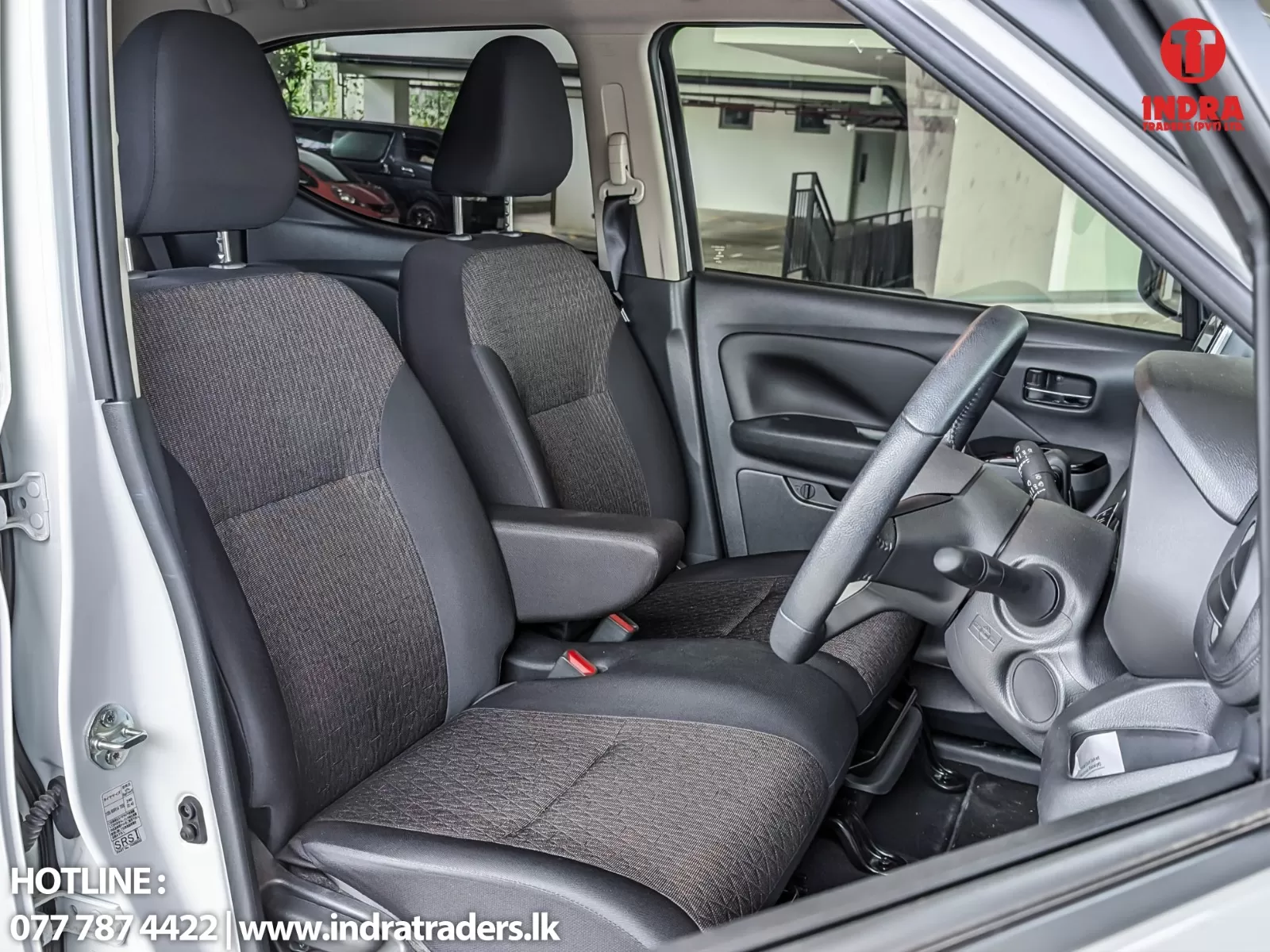 Interior of a right-hand-drive car: front seats, steering wheel, and dashboard visible from the open driver's door; showroom backdrop in background.
