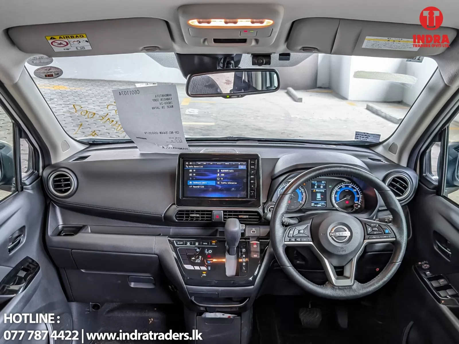 Interior view of a car cockpit from the driver’s seat, showing steering wheel, center touchscreen, gear lever, rearview mirror, and a sheet on the windshield with hotline text in the corner.