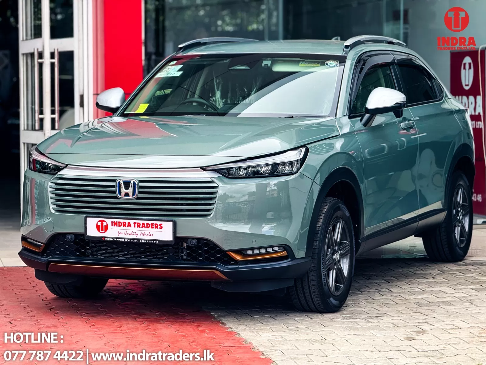 Mint-green Honda SUV shown from the front-right at a dealership, with Indra Traders branding on the plate area and storefront in the background.