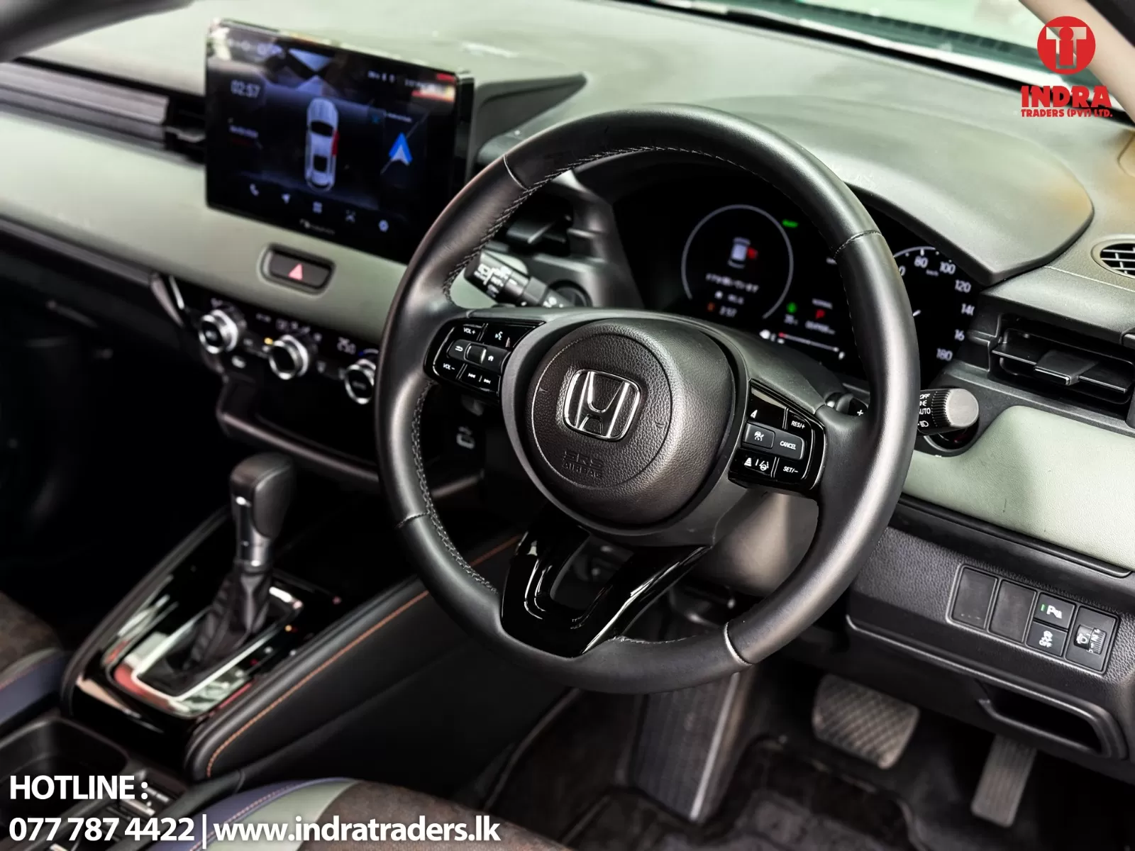 Interior of a Honda car showcasing the steering wheel, digital gauge cluster, and center touchscreen with gear shifter in view; Indra Traders logo in the top-right and contact text at bottom-left.