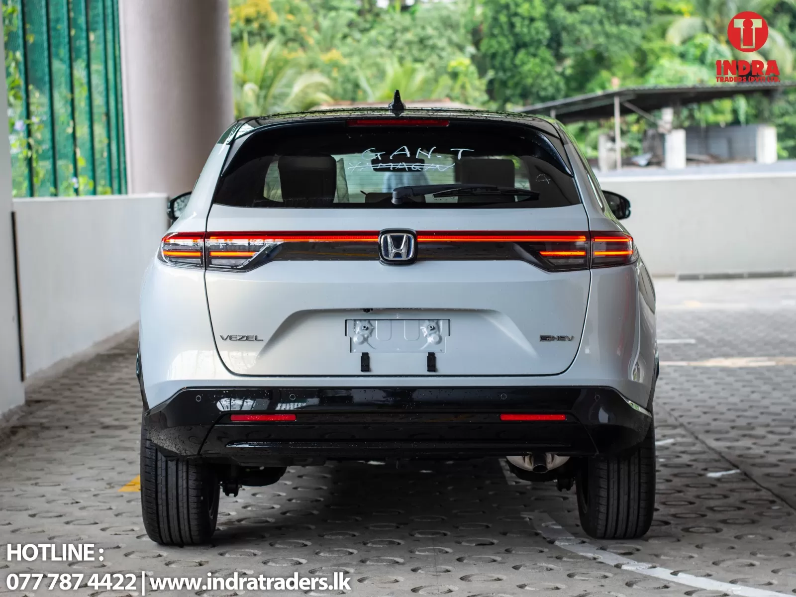 Rear view of a white Honda Vezel SUV in a parking area, bright taillights across the tailgate, Indra Traders logo in the top-right corner, hotline info at bottom-left.