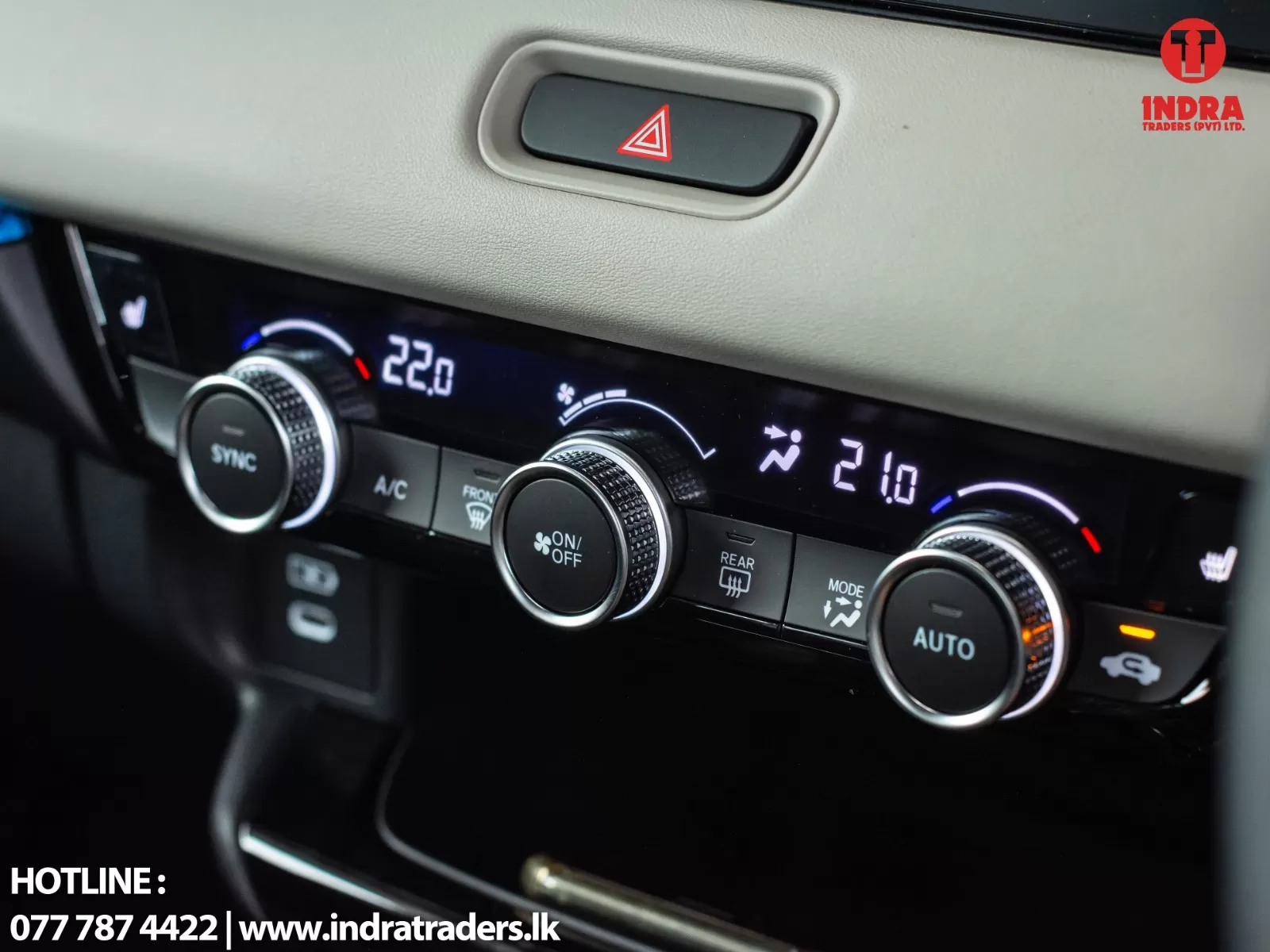 Close-up of a car's climate control panel showing dual temperatures (left 22.0°C, right 21.0°C) with a hazard button above and glossy dials on the control strip. Includes Indra Traders logo in the top-right corner.