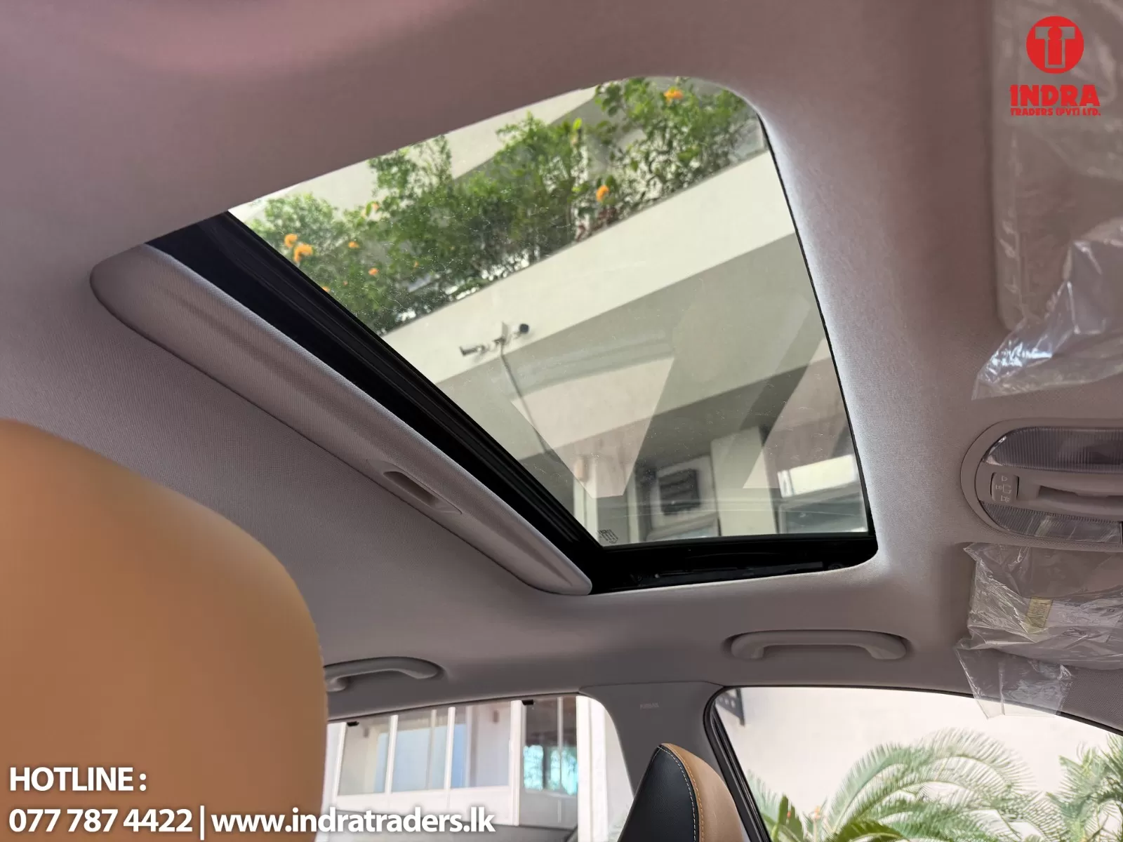 Interior of a car showing a panoramic sunroof with trees outside and a building facade visible through the glass, beige headliner and seats visible.
