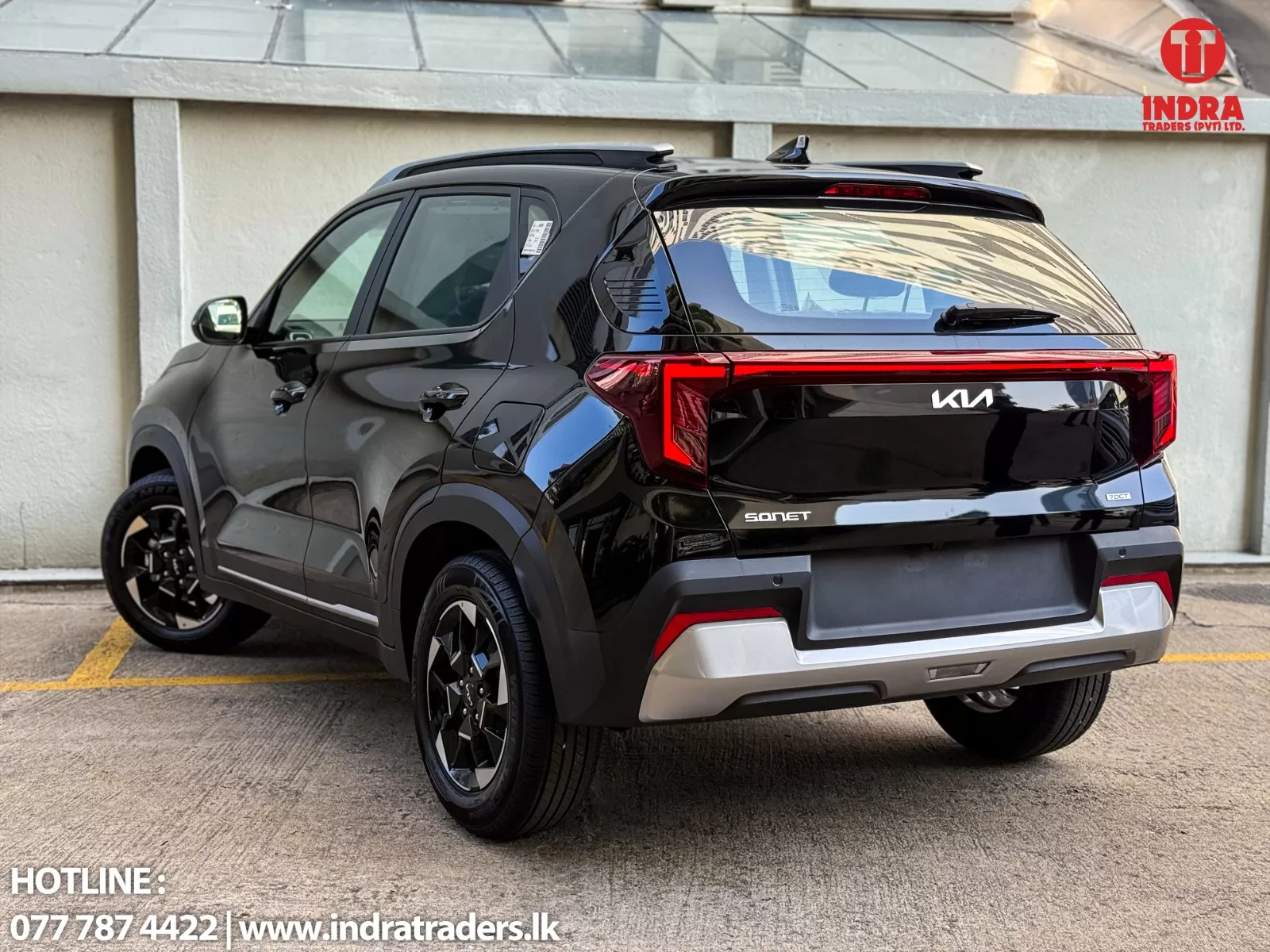 Black Kia Sonet SUV seen from rear-right, parked in lot with Indra Traders logo in corner, reflecting surrounding buildings in glossy surface.