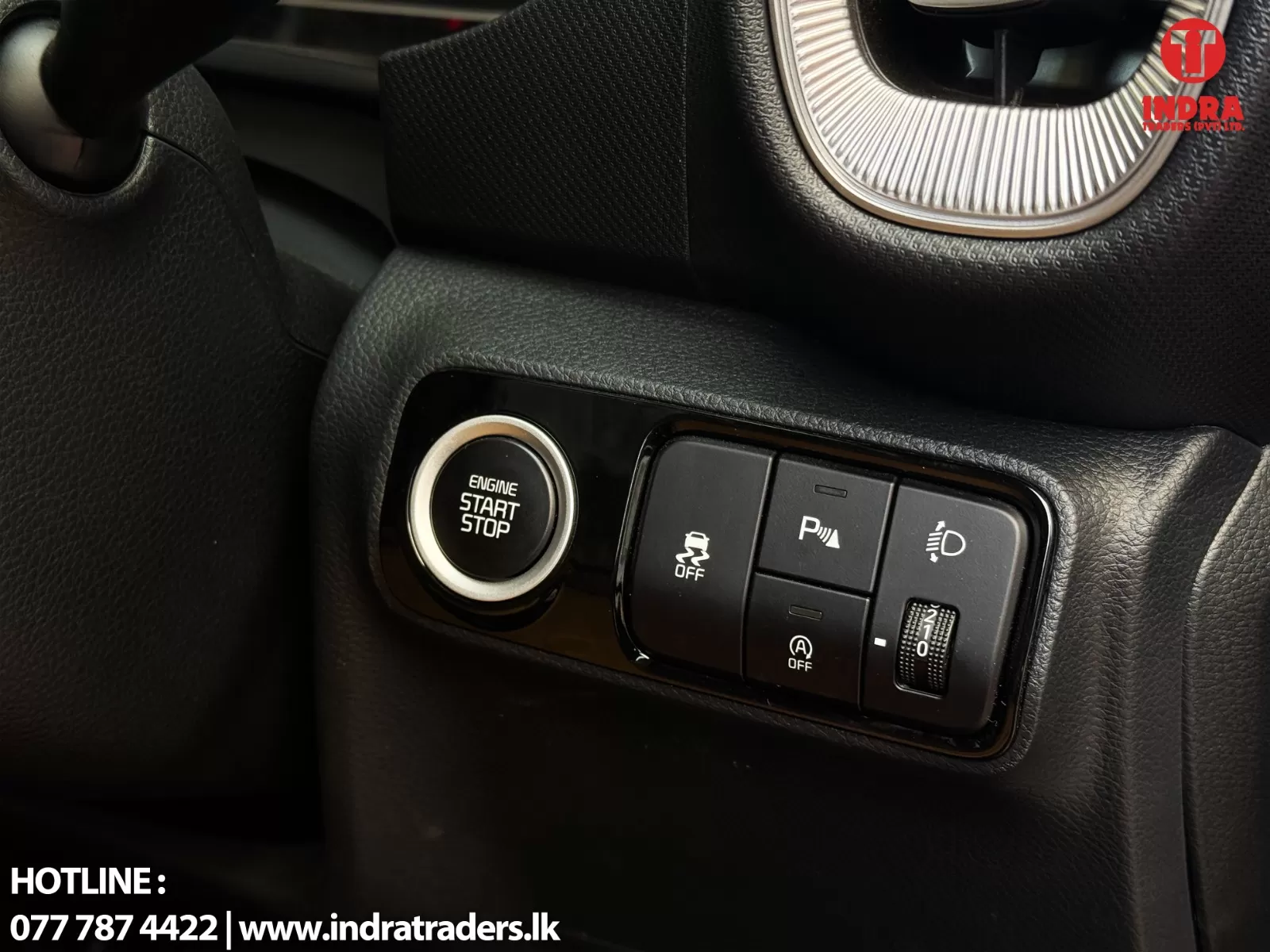 Car interior panel with engine start/stop button and control switches (traction off, parking assist, headlight adjust) and Indra Traders watermark in corner.