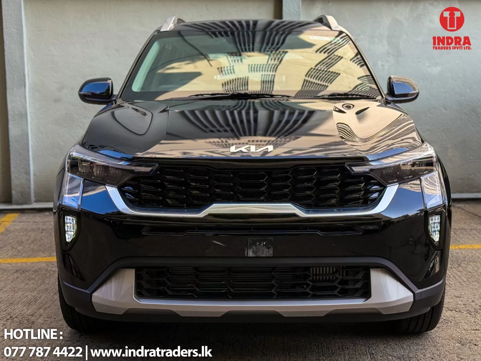 Front view of a glossy black Kia SUV with a bold grille, parked in a lot; Indra Traders logo in the upper-right corner and hotline/website text at the bottom.
