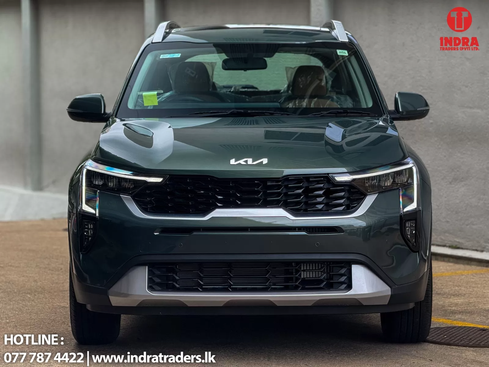 Front view of a dark green Kia SUV parked near a concrete wall, with the Indra Traders logo in the top-right corner and contact info along the bottom edge.
