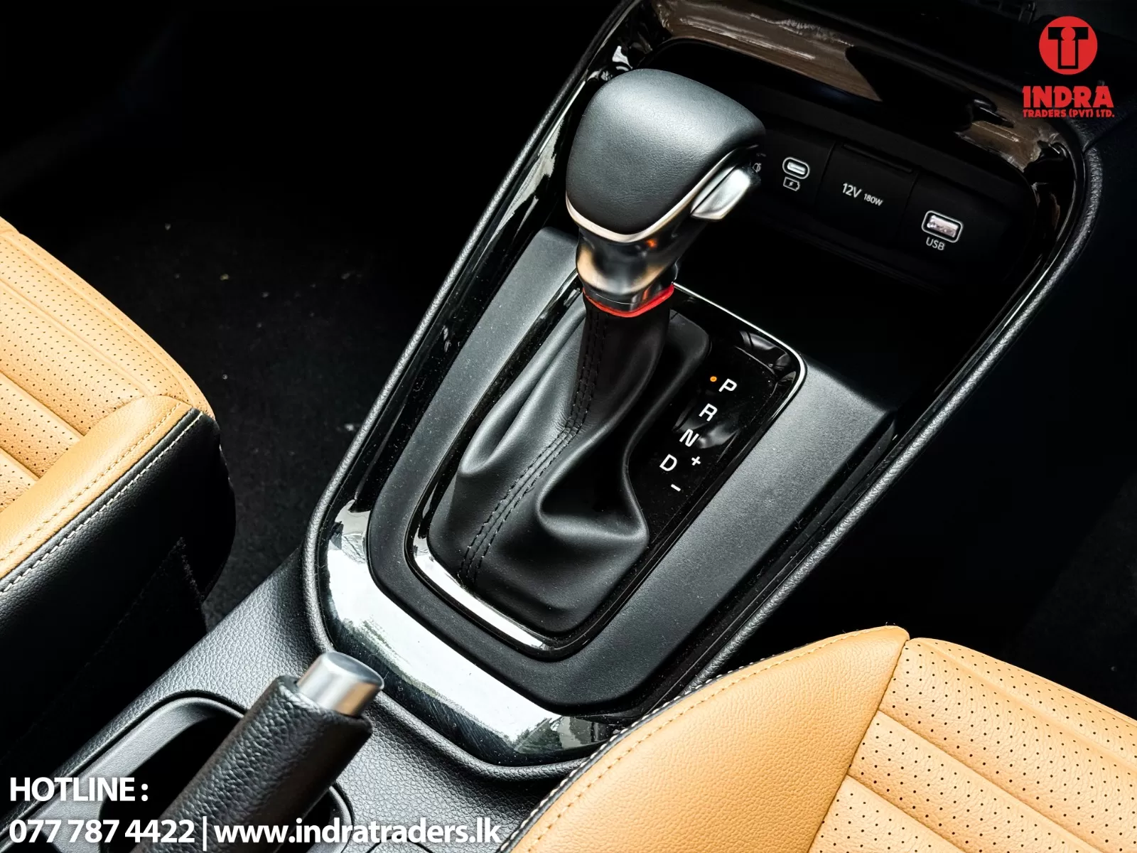 Close-up of a car’s automatic gear shift on a center console, with tan perforated seats and an Indra Traders watermark in the corner.