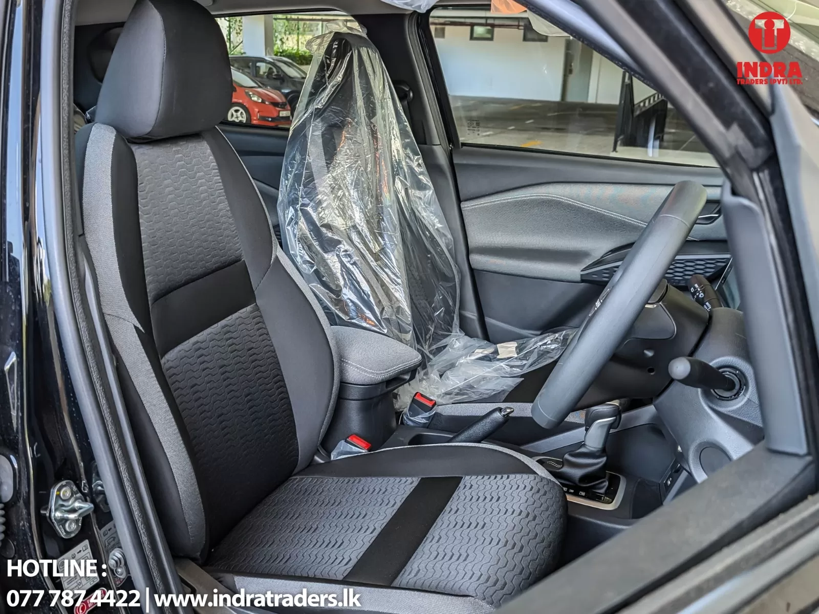Interior of a new car showing plastic wrapping on the driver seat, with the steering wheel, center console, and gray fabric seats visible in a showroom setting.