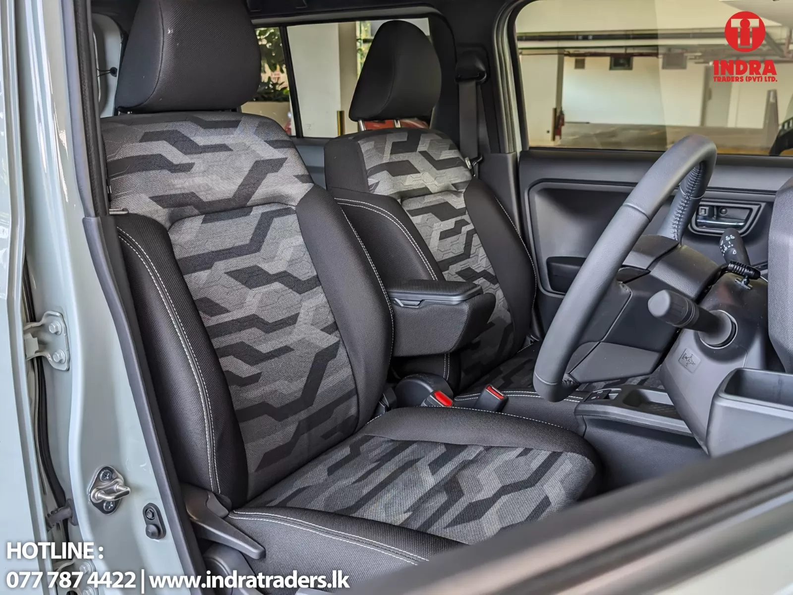 Interior of a car showing front seats with gray geometric-patterned upholstery and a center armrest, steering wheel and dashboard visible on the right; logo and contact text appear in the upper-right and lower-left corners.