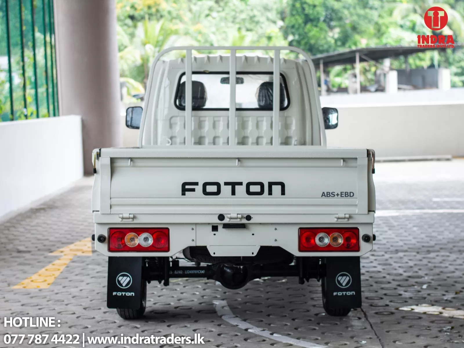 Rear view of a white FOTON pickup under a covered area; taillights visible and INDRA TRADERS logo appears in the top-right corner. Hotline and website text are shown at the bottom left.