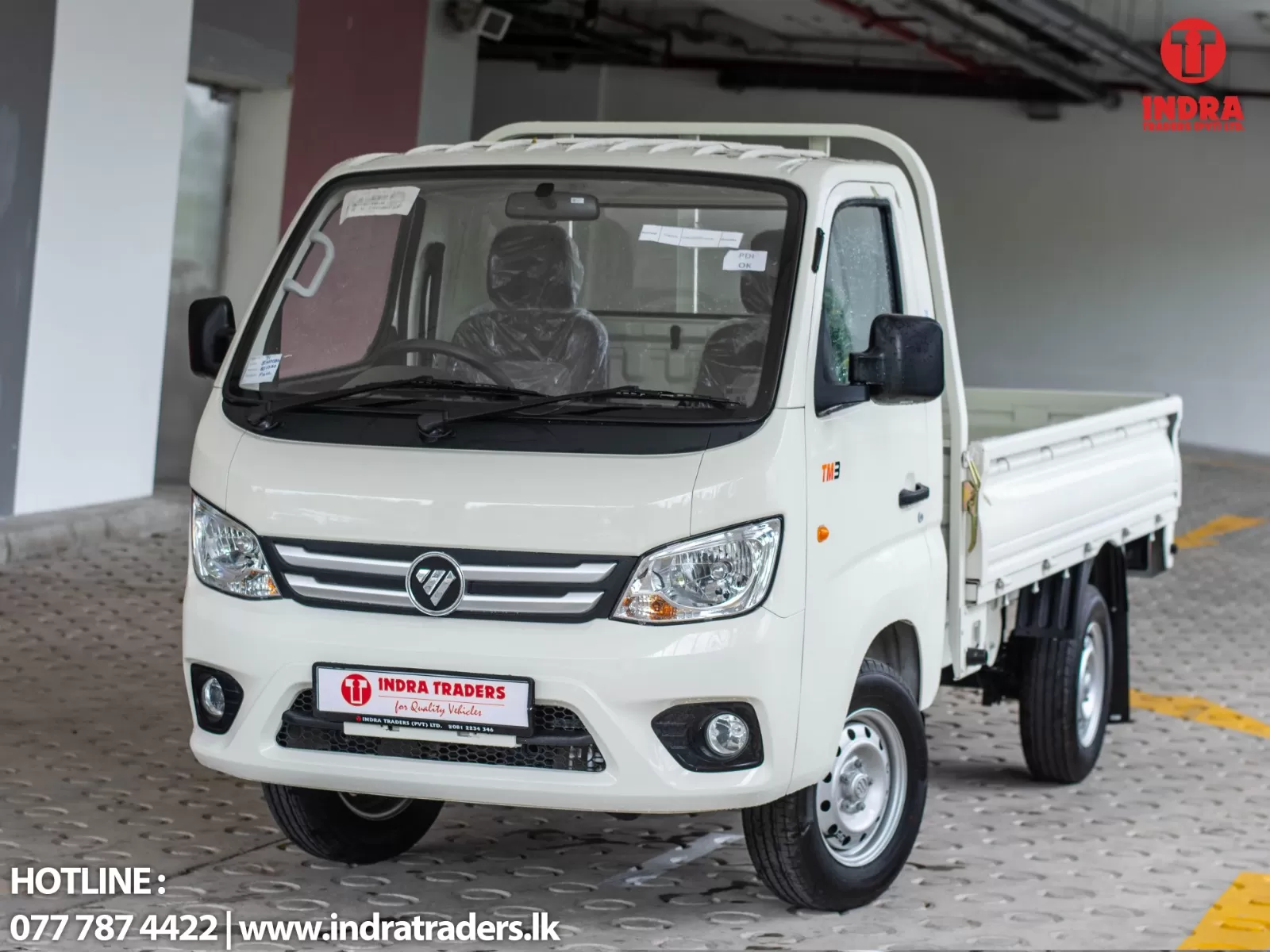 White flatbed truck shown in a showroom, front three-quarter view with windshield stickers and Indra Traders logo in the upper-right corner.