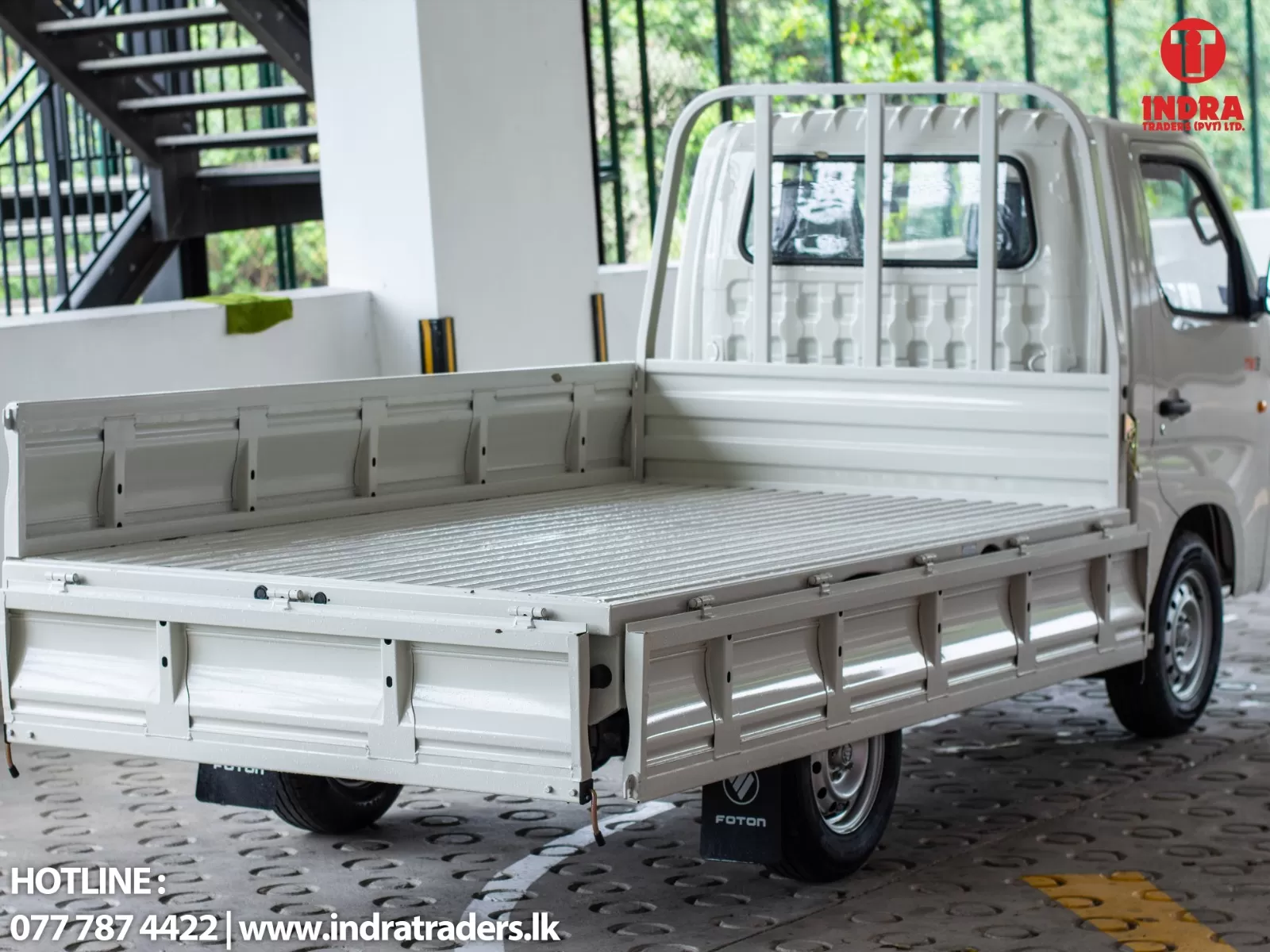 White pickup with an open flatbed in a showroom setting; Indra Traders logo in the top-right and hotline text at lower-left.
