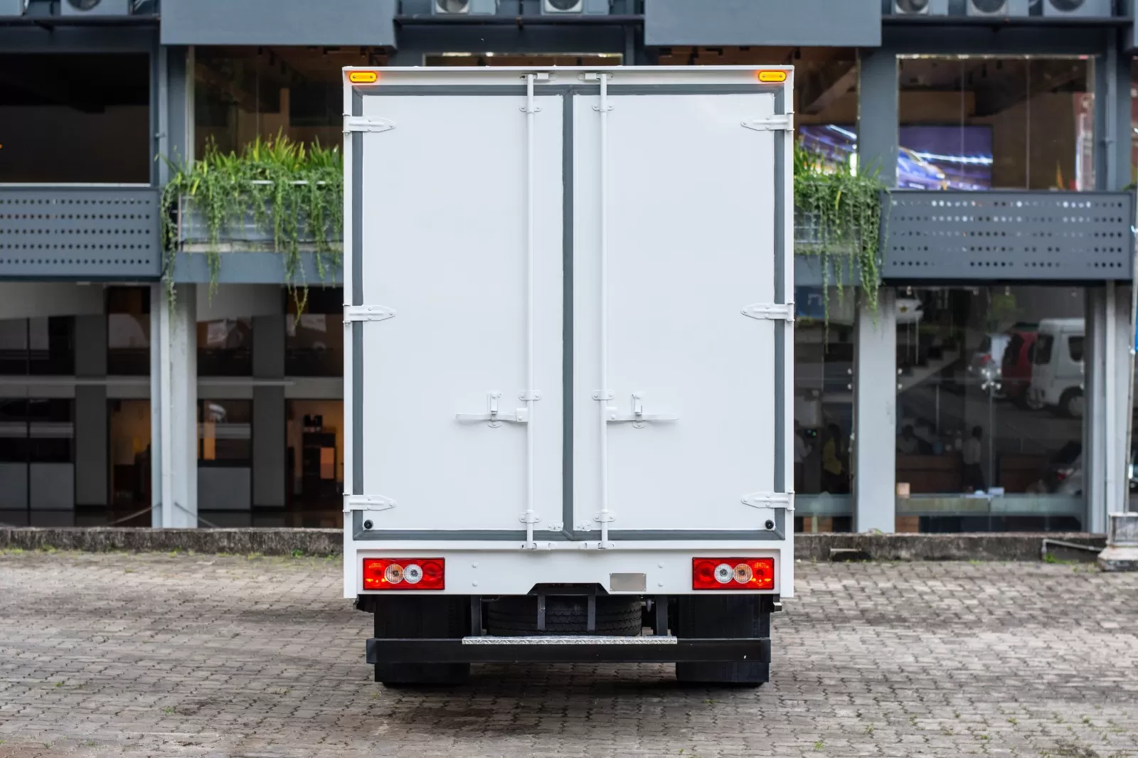 Rear view of a white delivery truck with closed double doors on a brick-paved lot, taillights lit and urban building in the background.