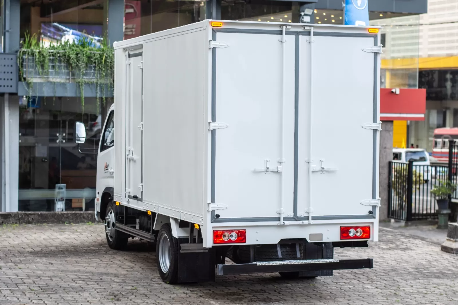 White delivery box truck parked on a cobblestone street, showing rear double doors and taillights in an urban setting
