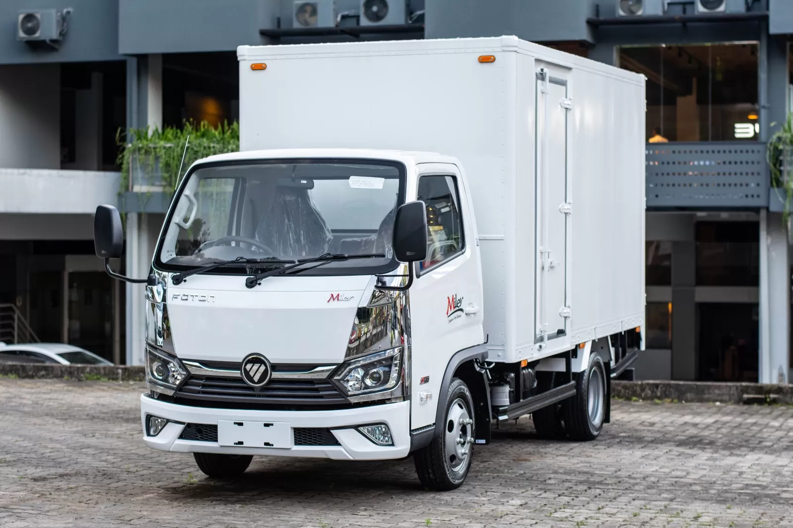 White delivery box truck parked on a cobblestone surface in an urban setting, front and side visible with company logo on door.