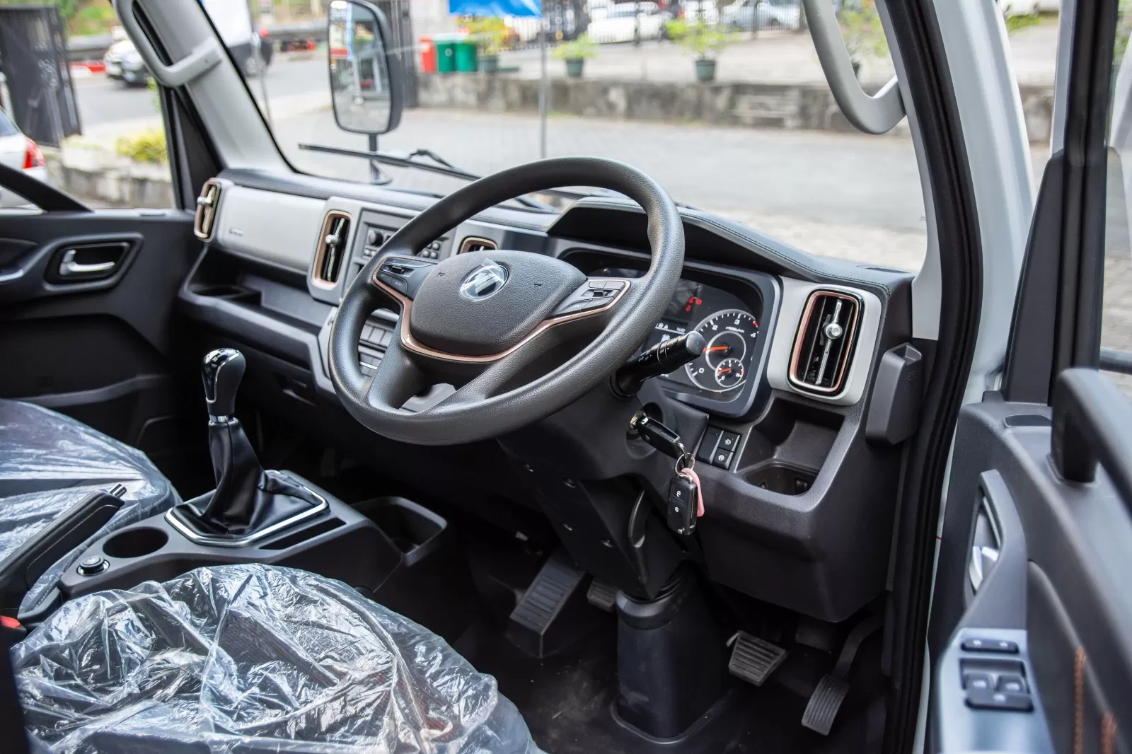 Interior of a vehicle cabin featuring a right-hand drive steering wheel, dashboard gauges, and a manual gear stick with seat covered in plastic wrap.