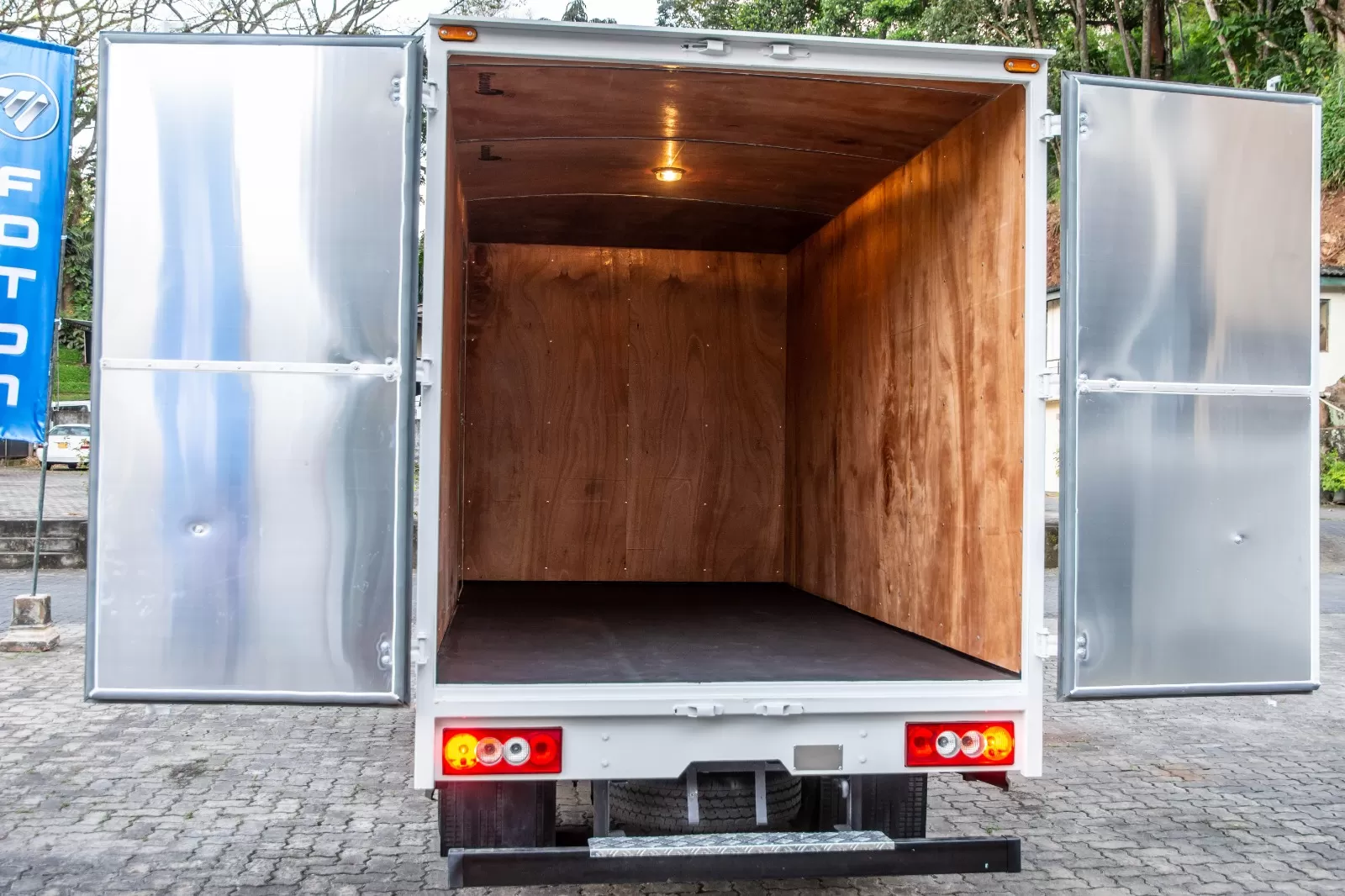 Open rear of a white cargo truck with plywood interior, doors fully swung open onto a paved area.