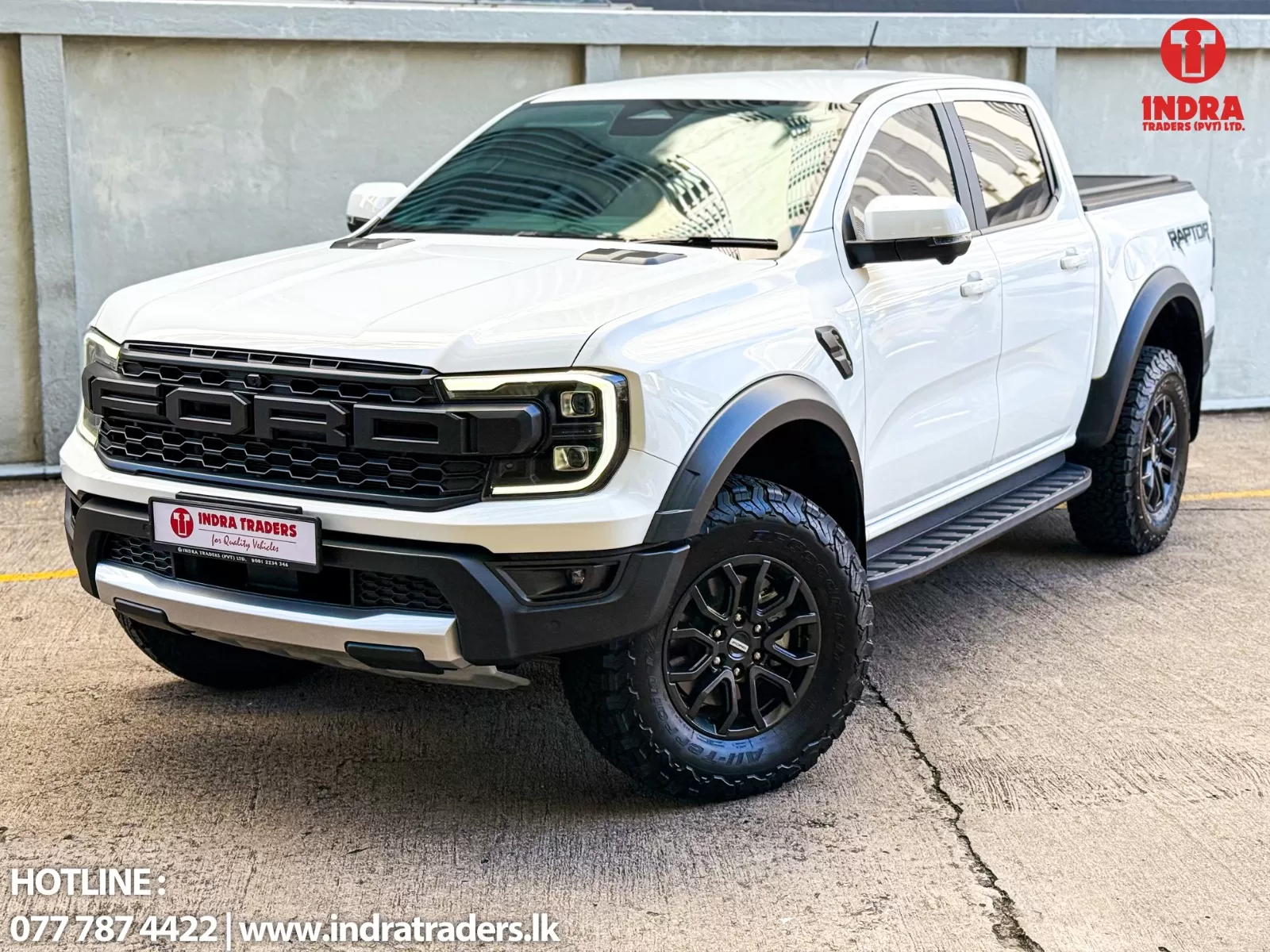 White Ford pickup with rugged tires parked in front of a concrete wall; Indra Traders branding appears on the top-right corner and license plate.