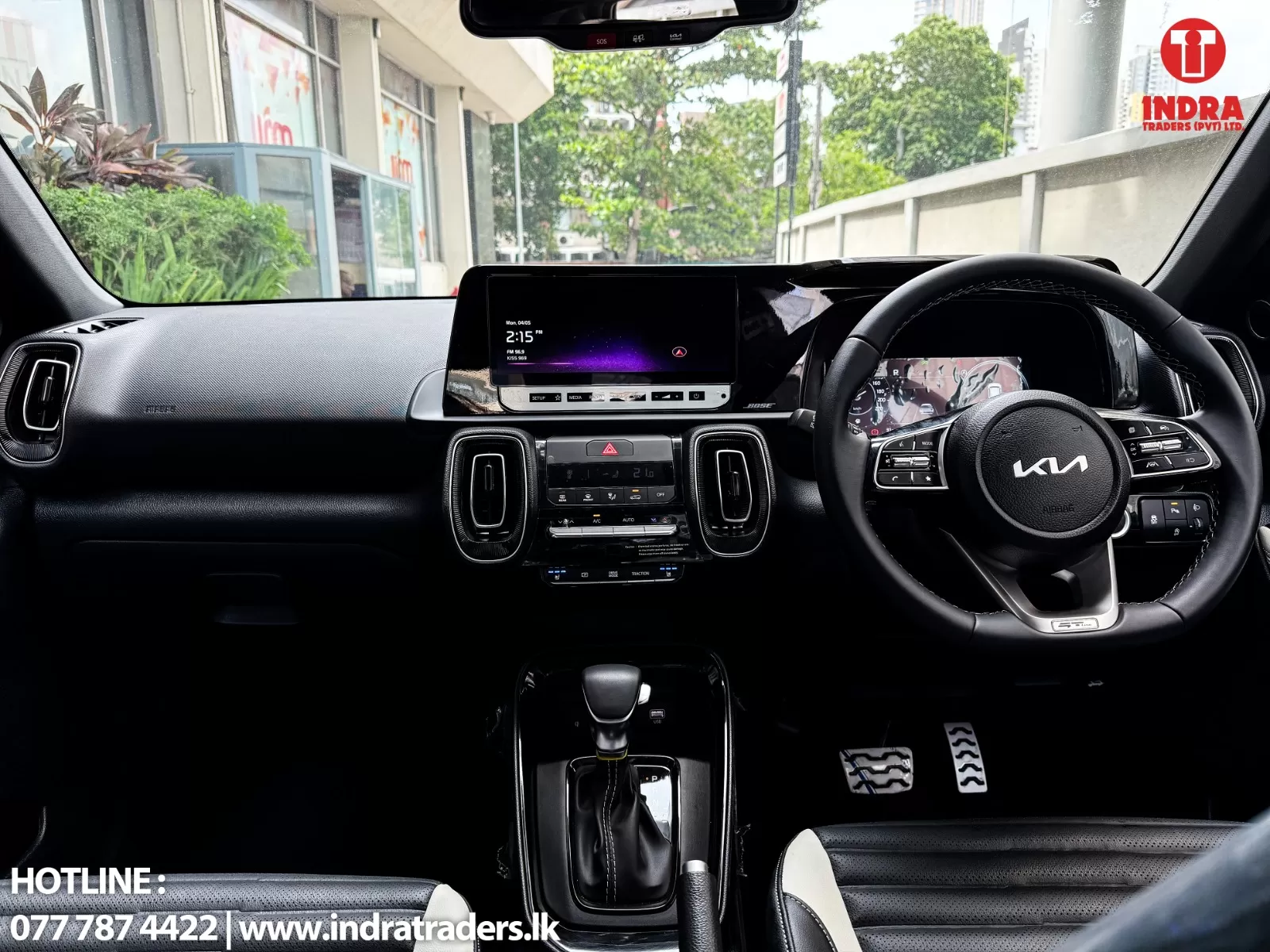 Interior view of a Kia car dashboard: steering wheel on the right, large central touchscreen, climate controls, and gear shifter