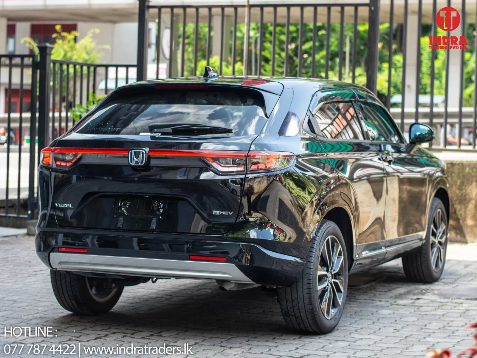Rear three-quarter view of a shiny black Honda Vezel e:HEV SUV parked near a metal fence; dealership watermark in the top-right corner.